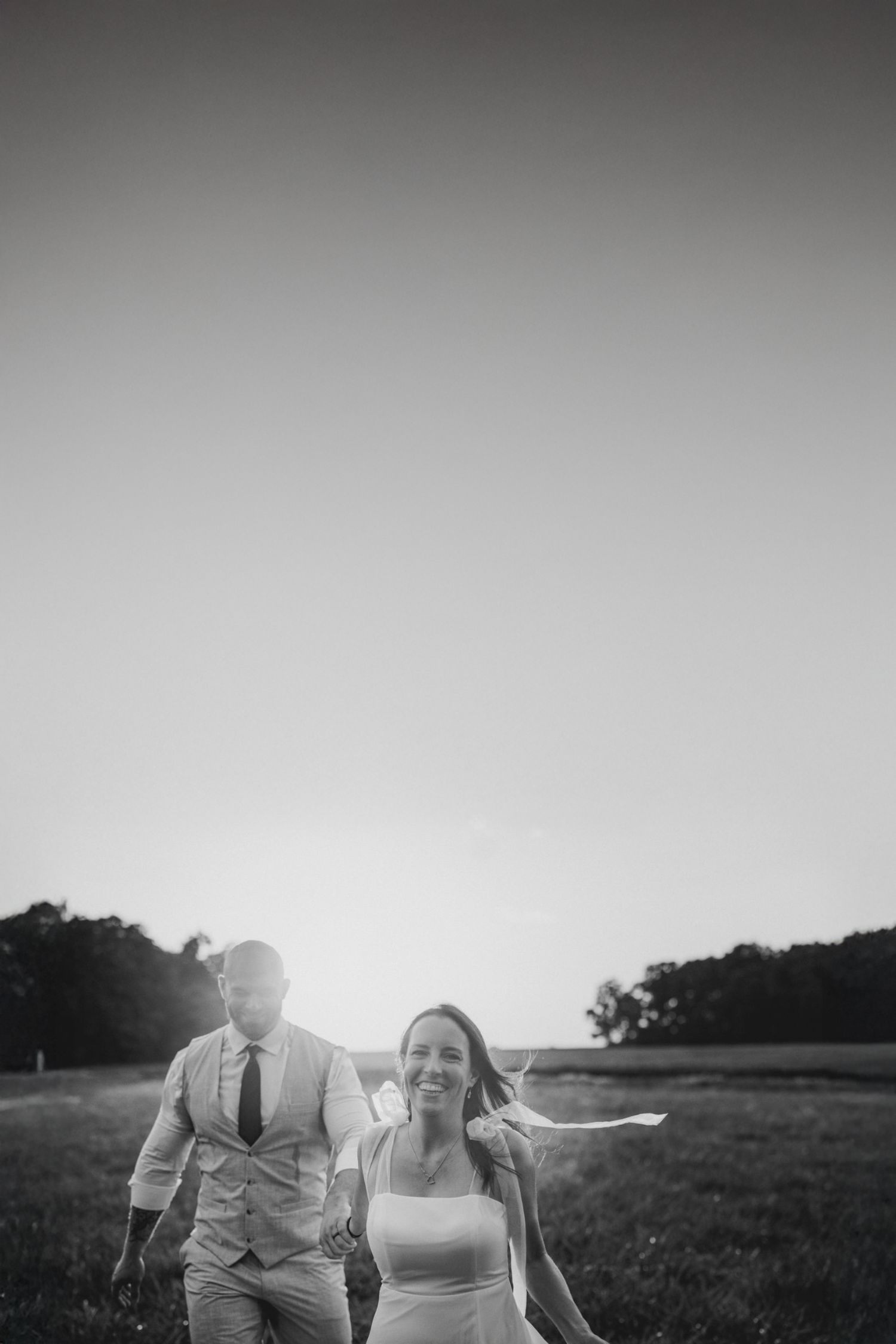 Black and white photo of a couple walking together in a field at sunset with a joyful expression.