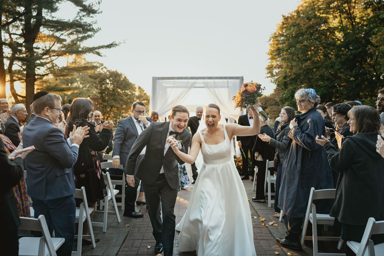 Outdoor wedding ceremony with guests seated in white chairs as golden sunlight filters through pine trees.
