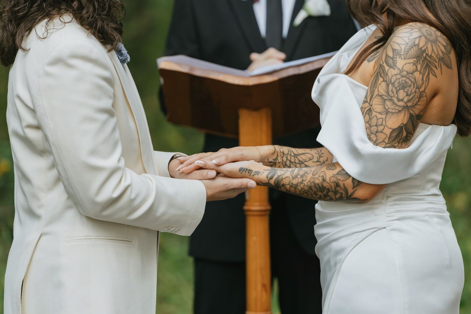 Two brides holding hands during outdoor wedding ceremony, one wearing white suit, other in backless gown with tattoos.