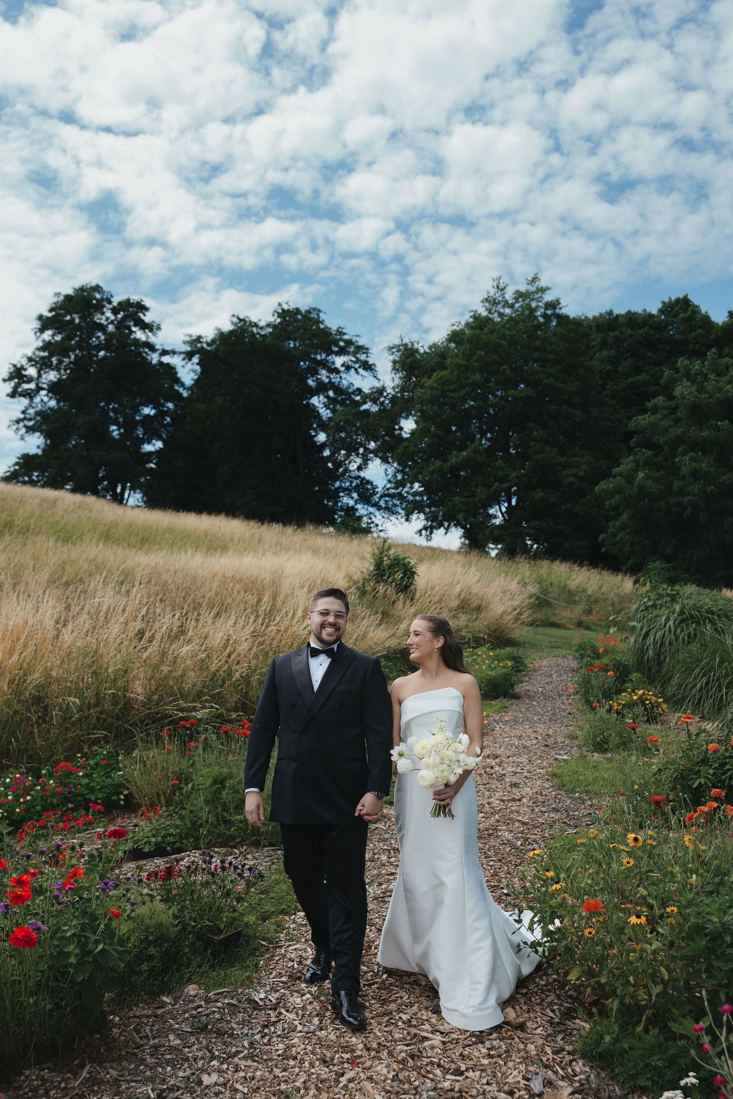 A bride and groom walk hand-in-hand through a flower garden path, she in a white gown, he in a black tuxedo.