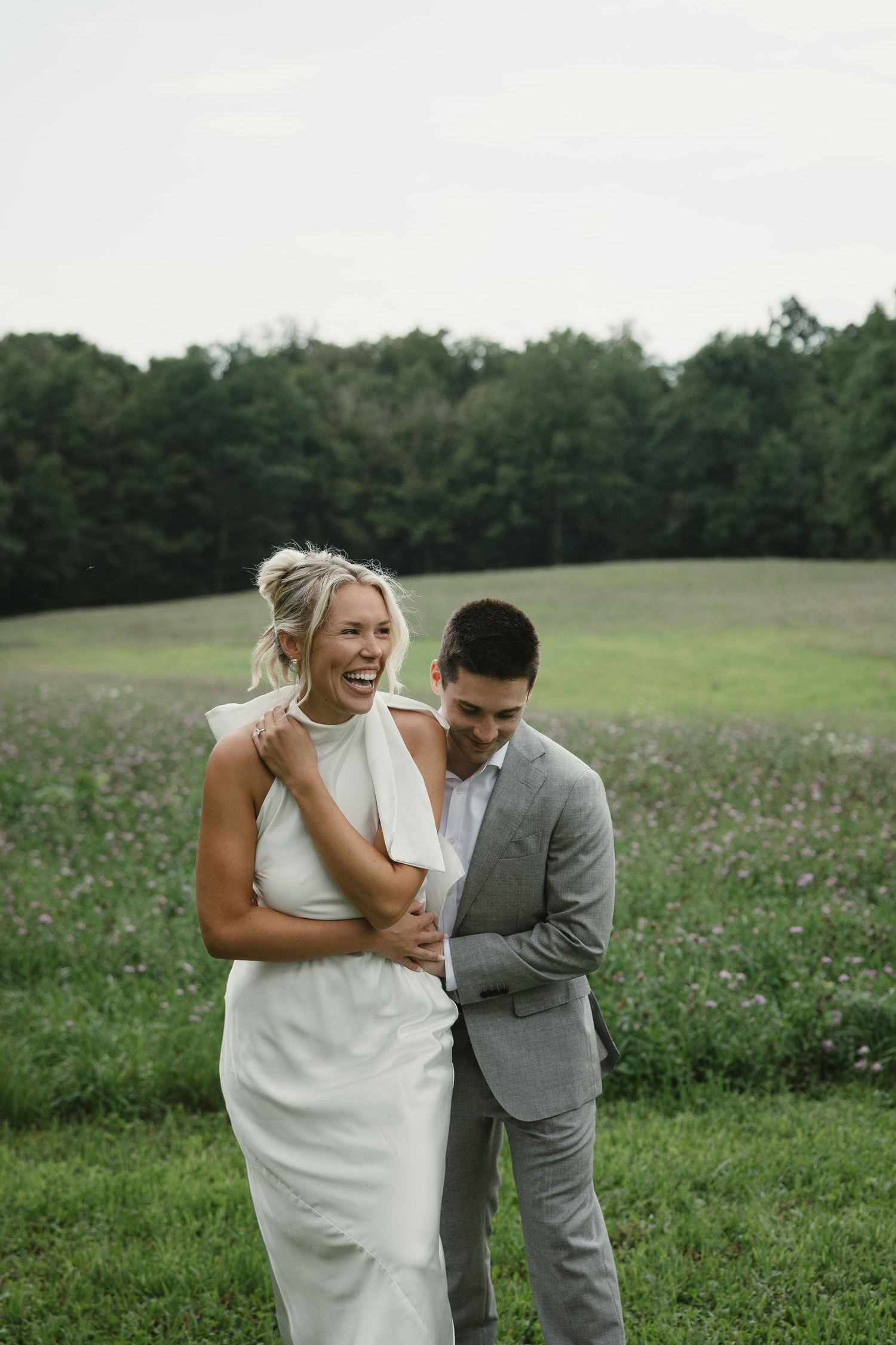 A laughing bride in a white dress and groom in a gray suit share a joyful moment in a lush green meadow.