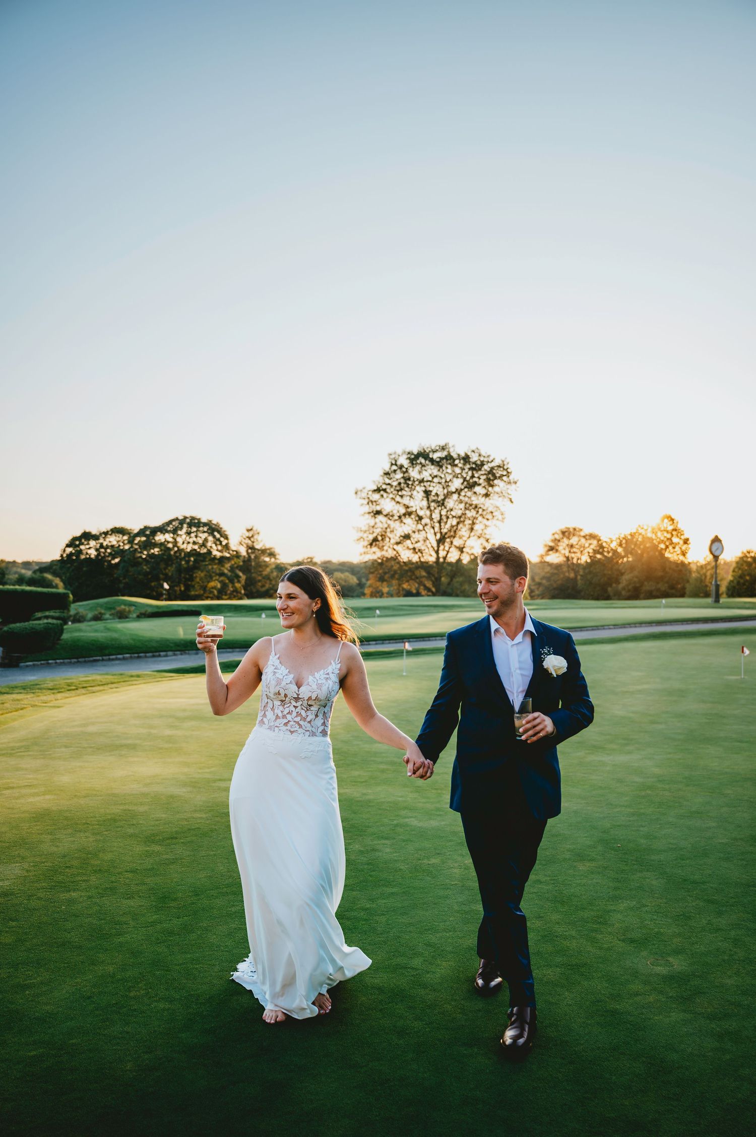 A couple walks hand in hand across a lush green golf course at sunset, creating a romantic silhouette.