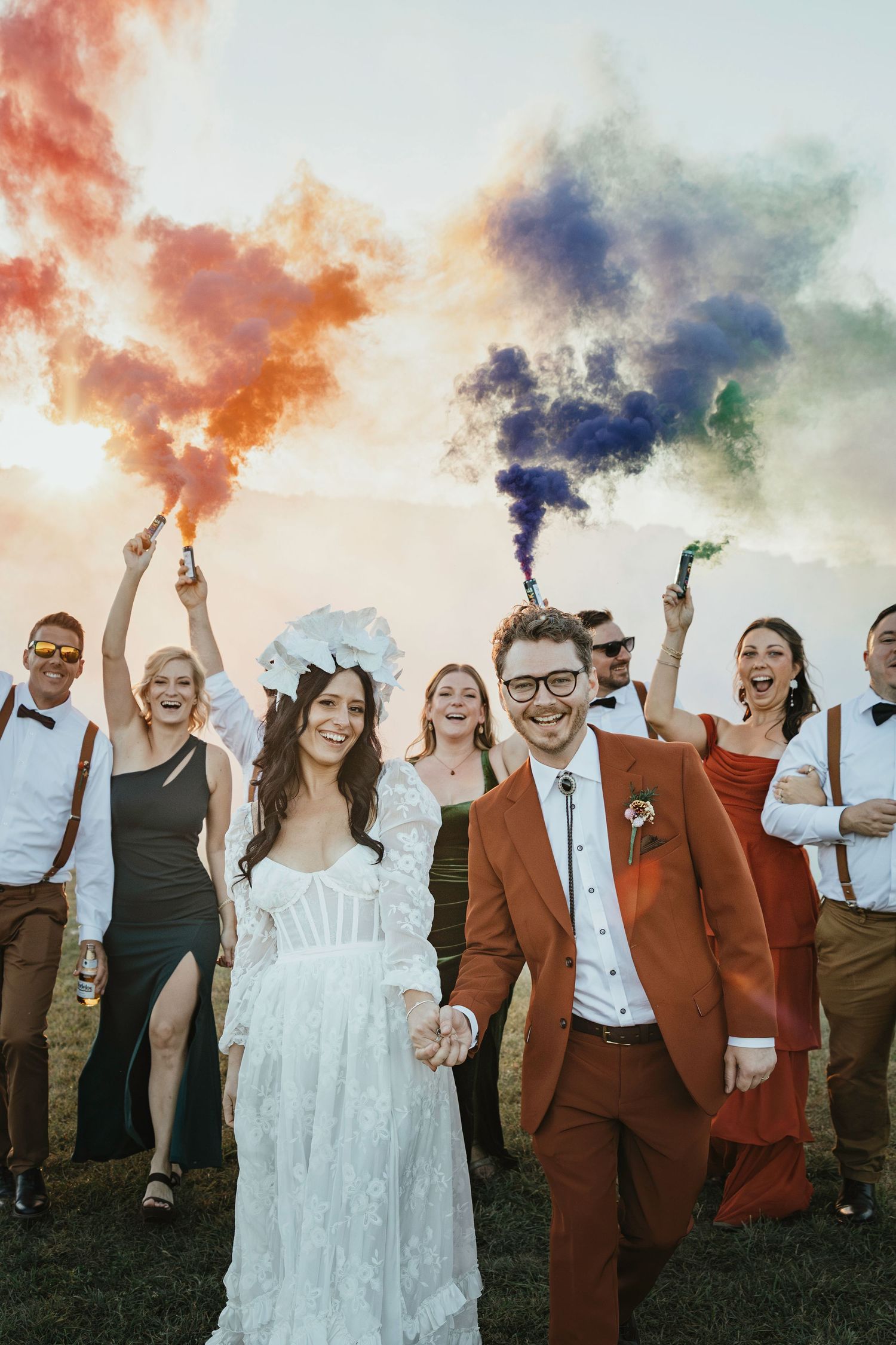 Joyful wedding party celebrating outdoors with colorful smoke bombs, bride in white gown and groom in rust suit holding hands.