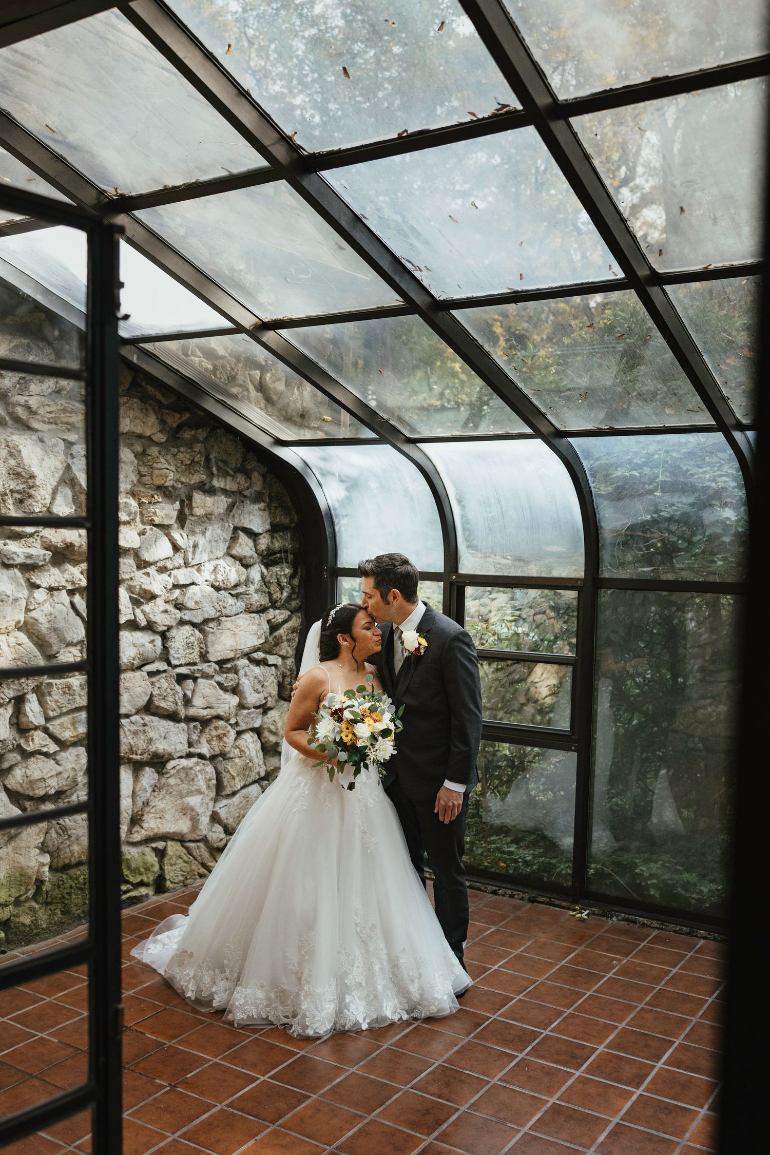 A couple sharing an intimate moment in a glass-enclosed conservatory with stone walls and terracotta flooring.