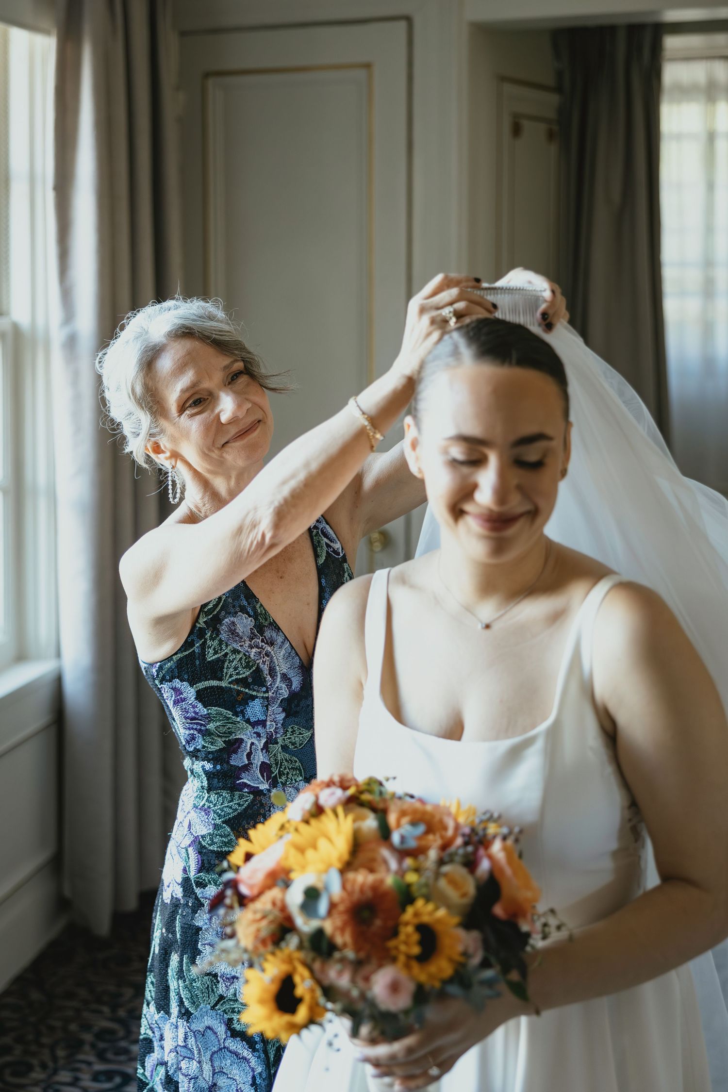 Bride and bridesmaid share joyful moments while getting ready in a bright room with sunflowers in the bouquet.