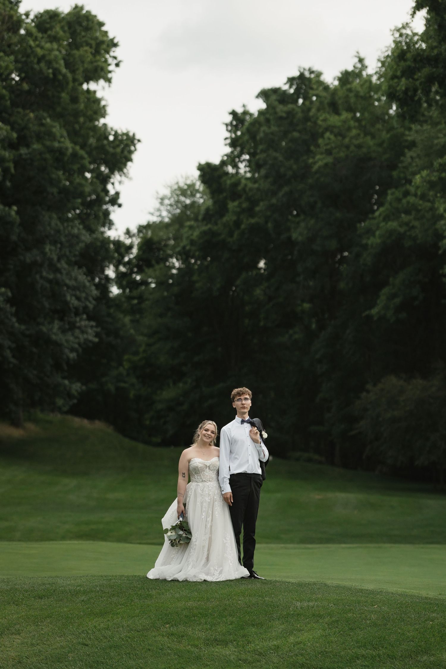 Bride in white gown and groom in black tuxedo standing on lush green golf course surrounded by trees.