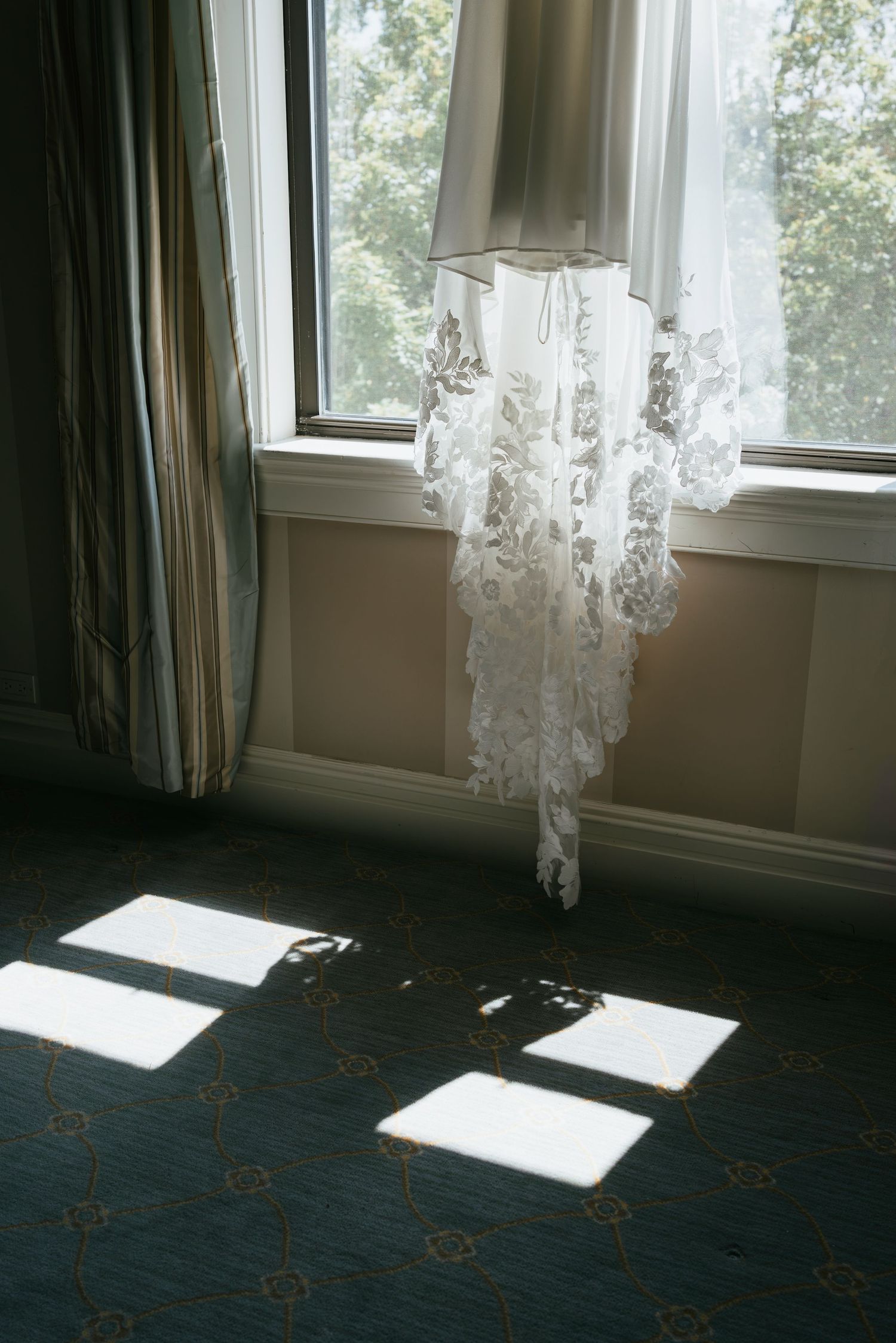 A delicate lace wedding veil hangs by a window, casting geometric shadows on a dark wooden floor.