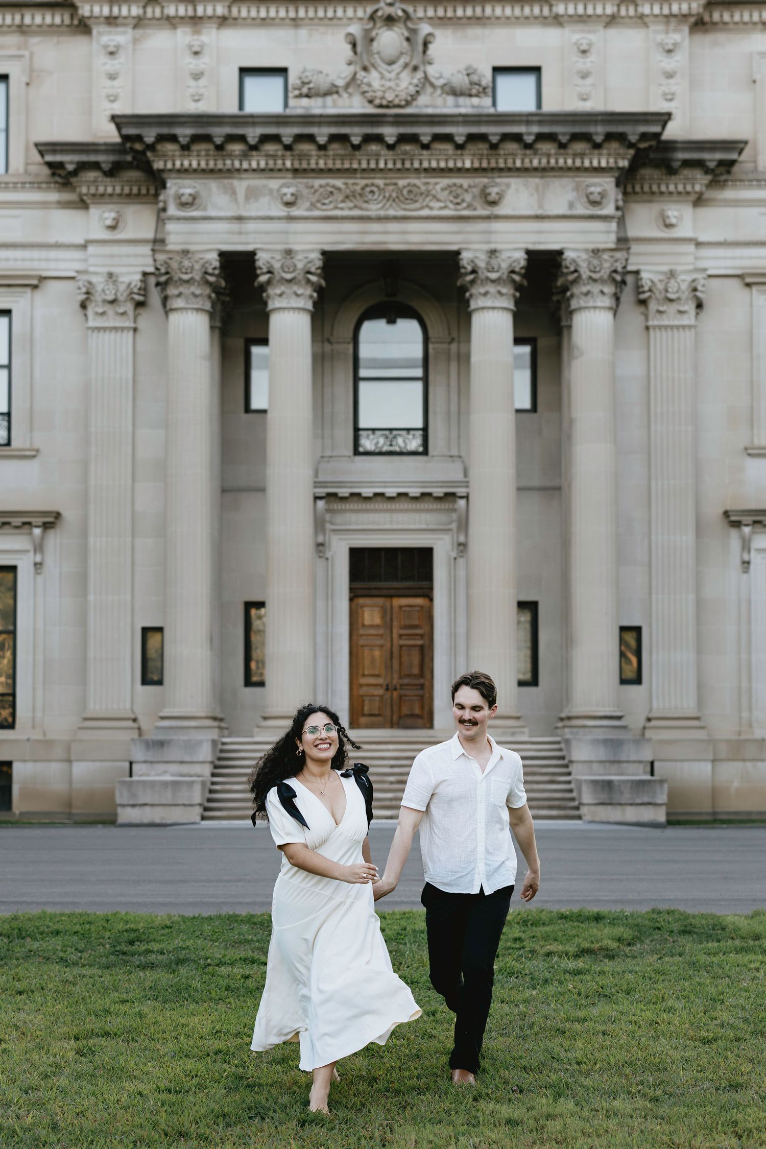 A couple in white outfits holds hands running on grass before a grand neoclassical stone building with columns.