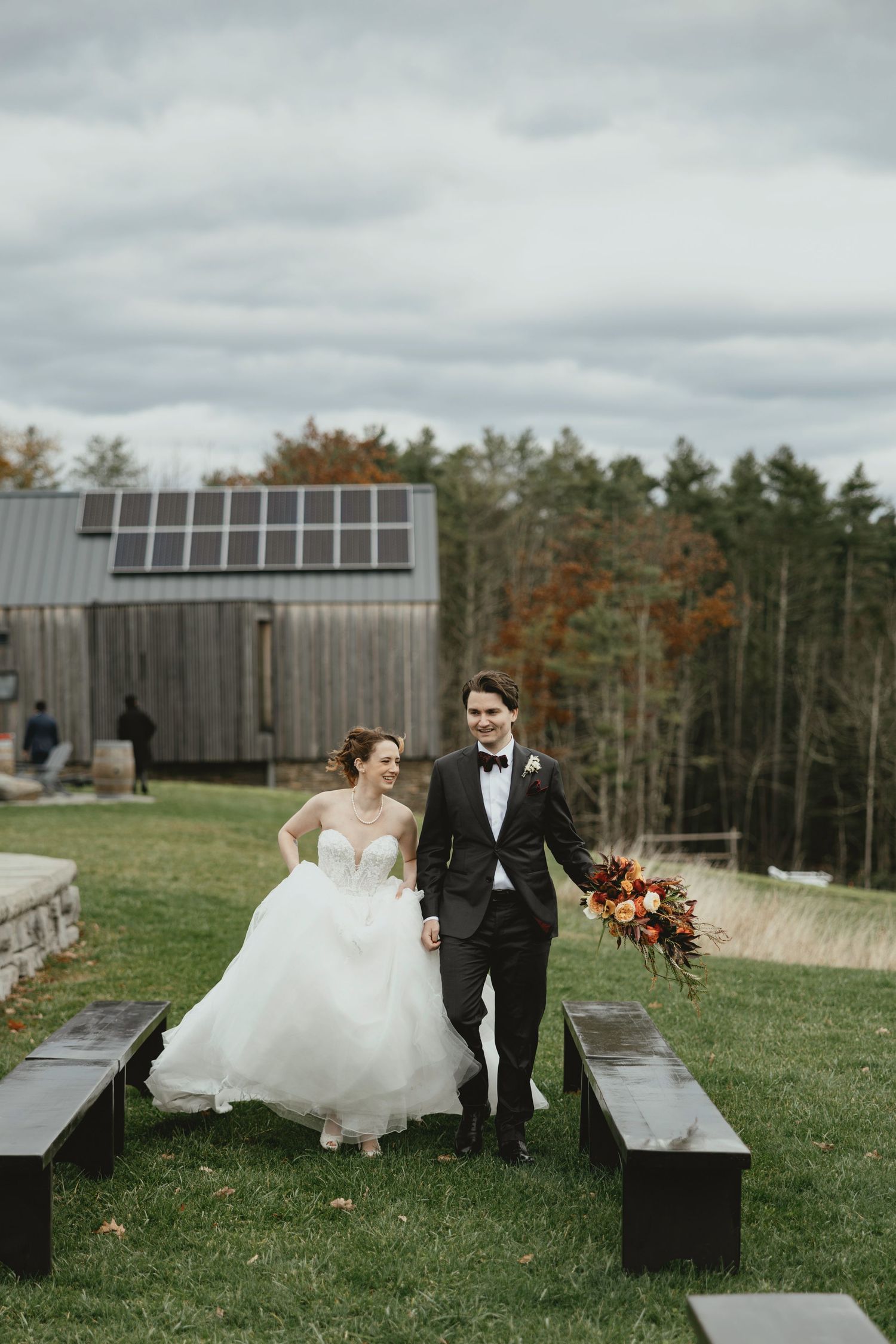 Wedding couple sitting on wooden benches at outdoor venue with autumn trees in background.