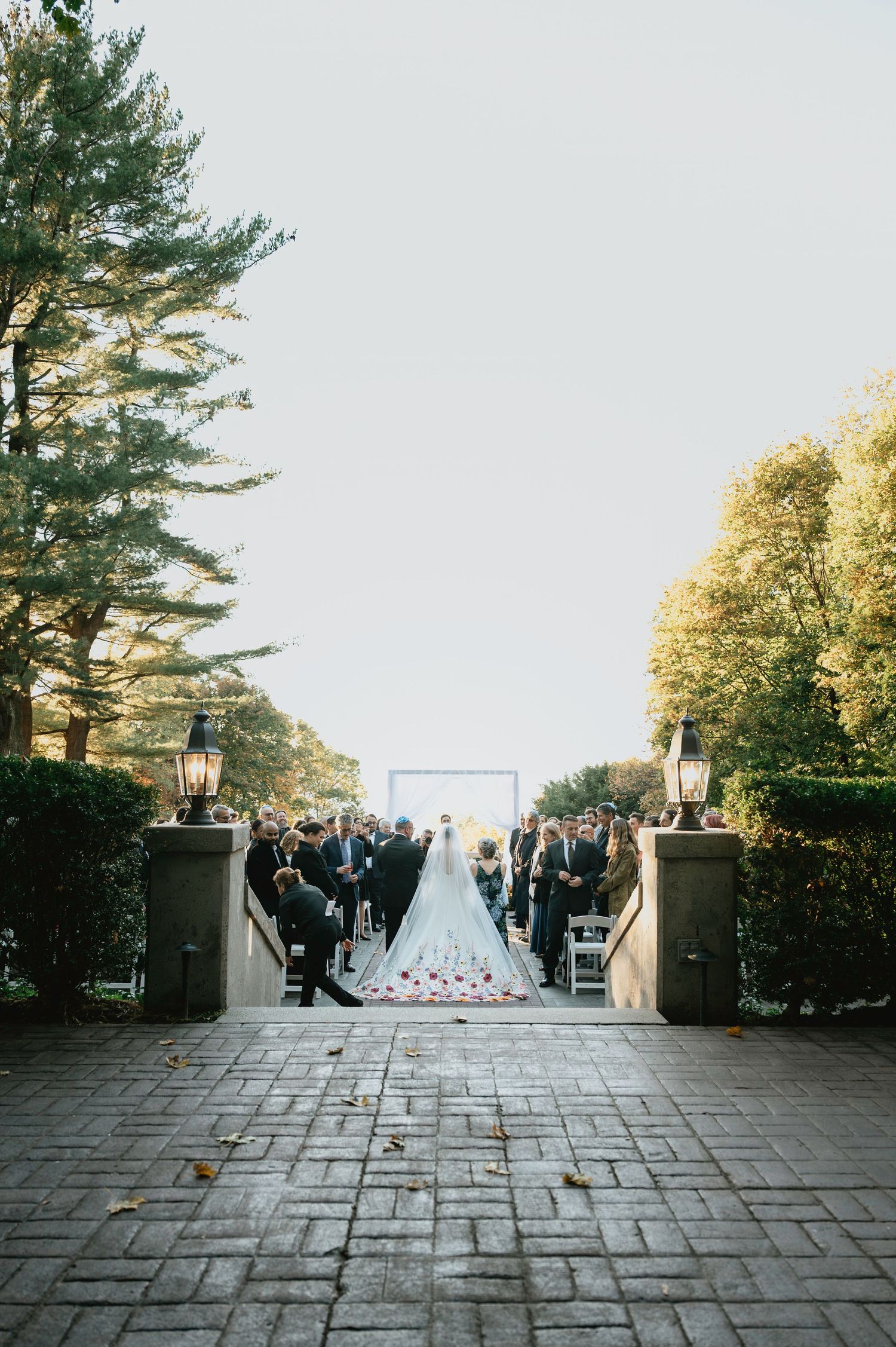 Wedding ceremony setup at an elegant outdoor stone patio venue with tall pine trees and lanterns at dusk.