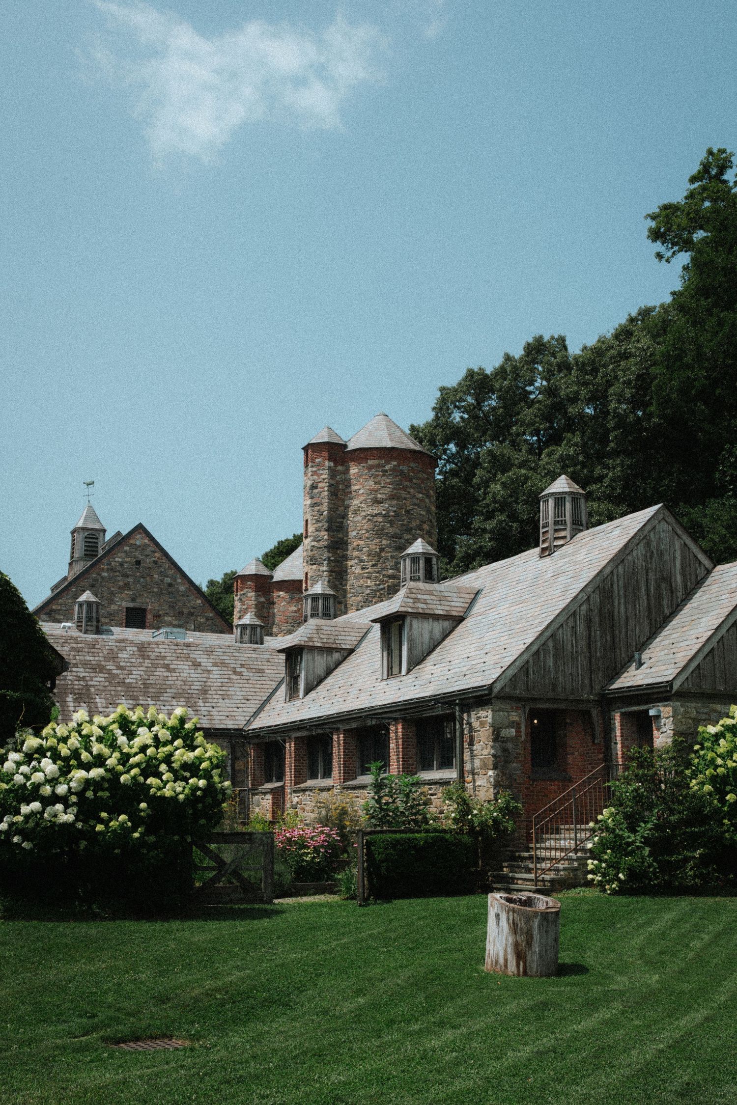 Historic stone farmhouse with gray slate roof, brick chimneys, white hydrangeas, and lush green lawn under clear blue sky.