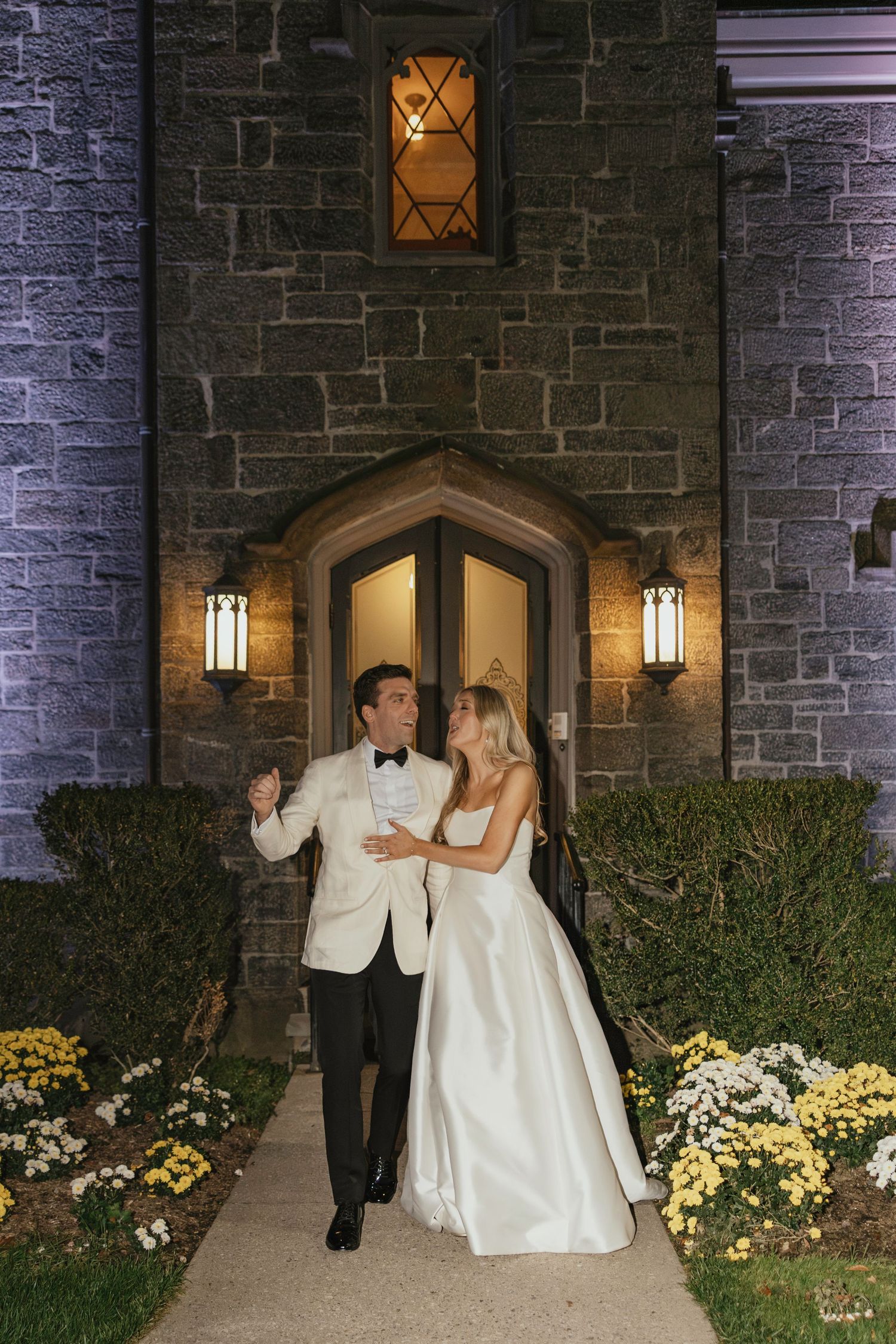 A wedding couple shares romantic moments in front of a stone church with illuminated doorways and yellow flowers.
