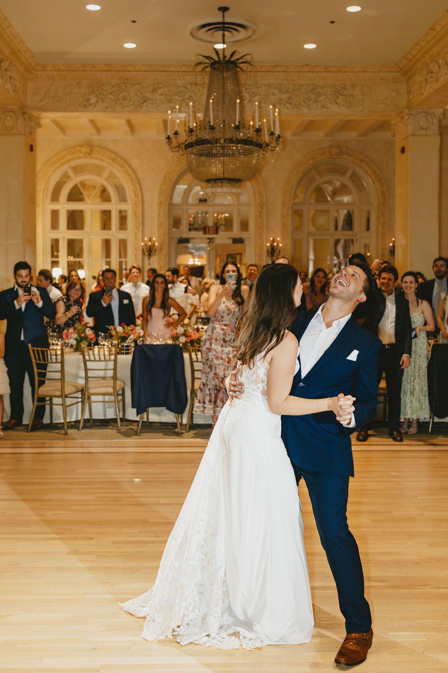 A couple shares their first dance in an elegant ballroom with ornate chandeliers and golden decor.