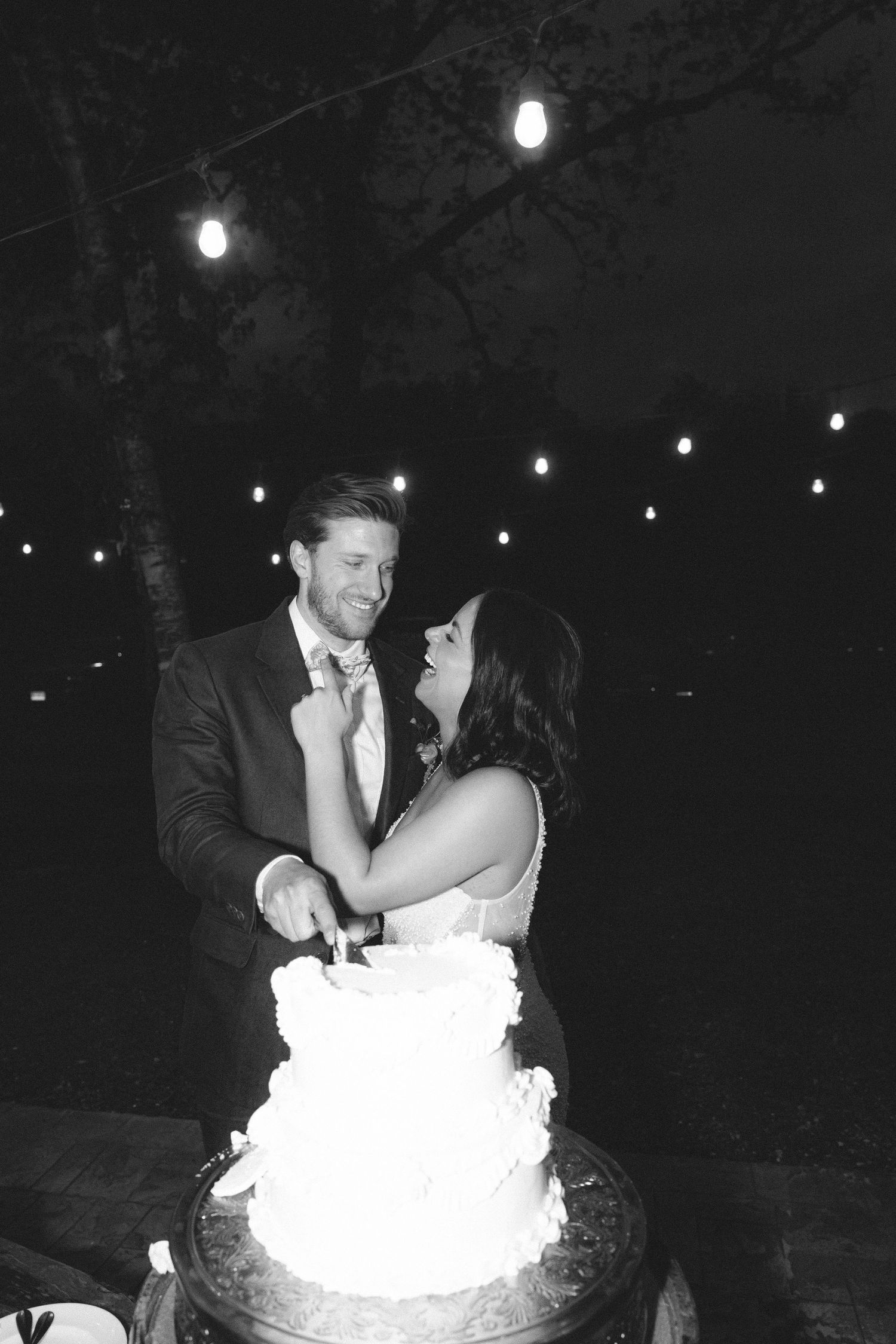 A romantic wedding couple shares their first slice of cake together at an evening outdoor reception with string lights.