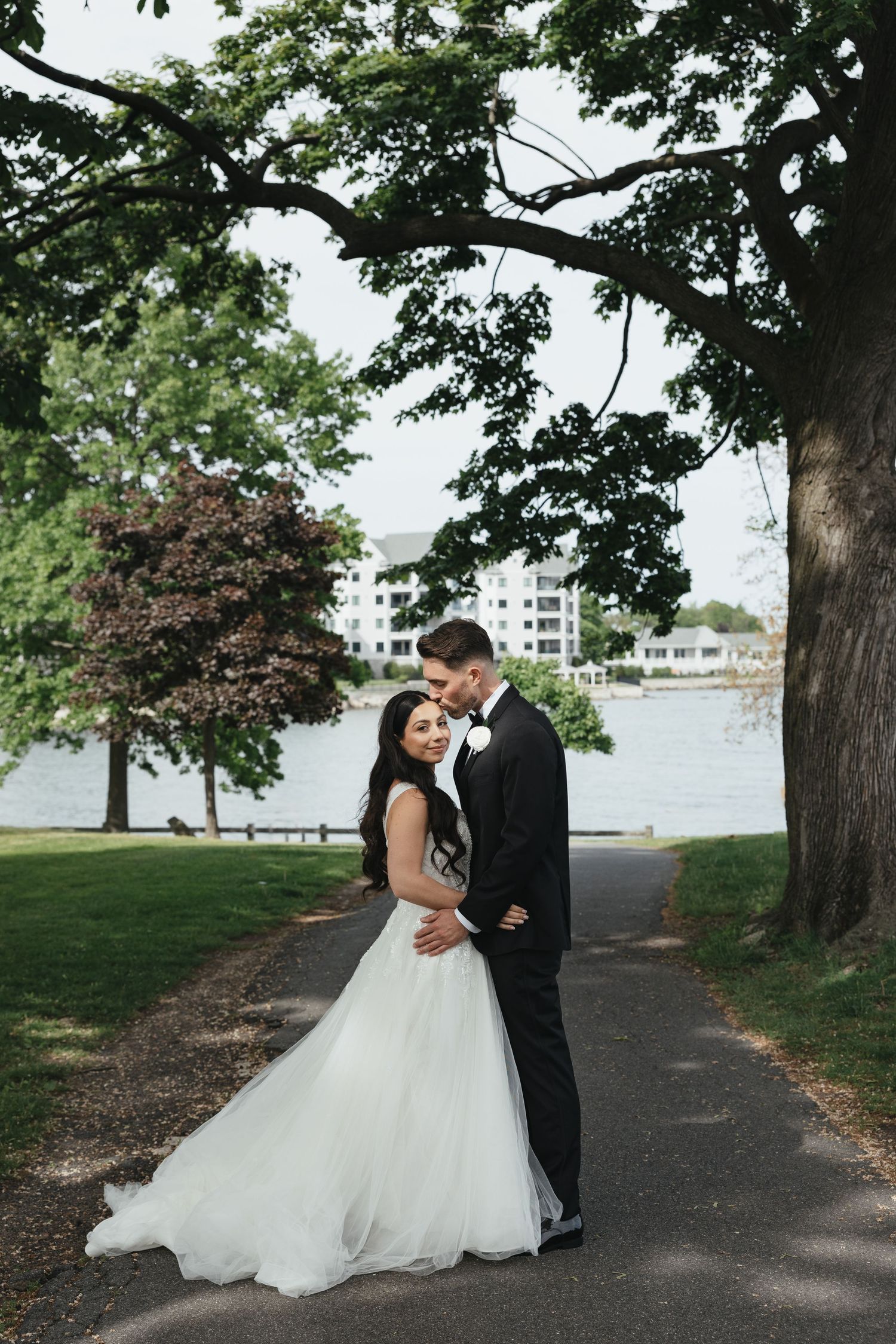 Bride in white gown and groom in black tuxedo embrace on a tree-lined path by a serene lake.