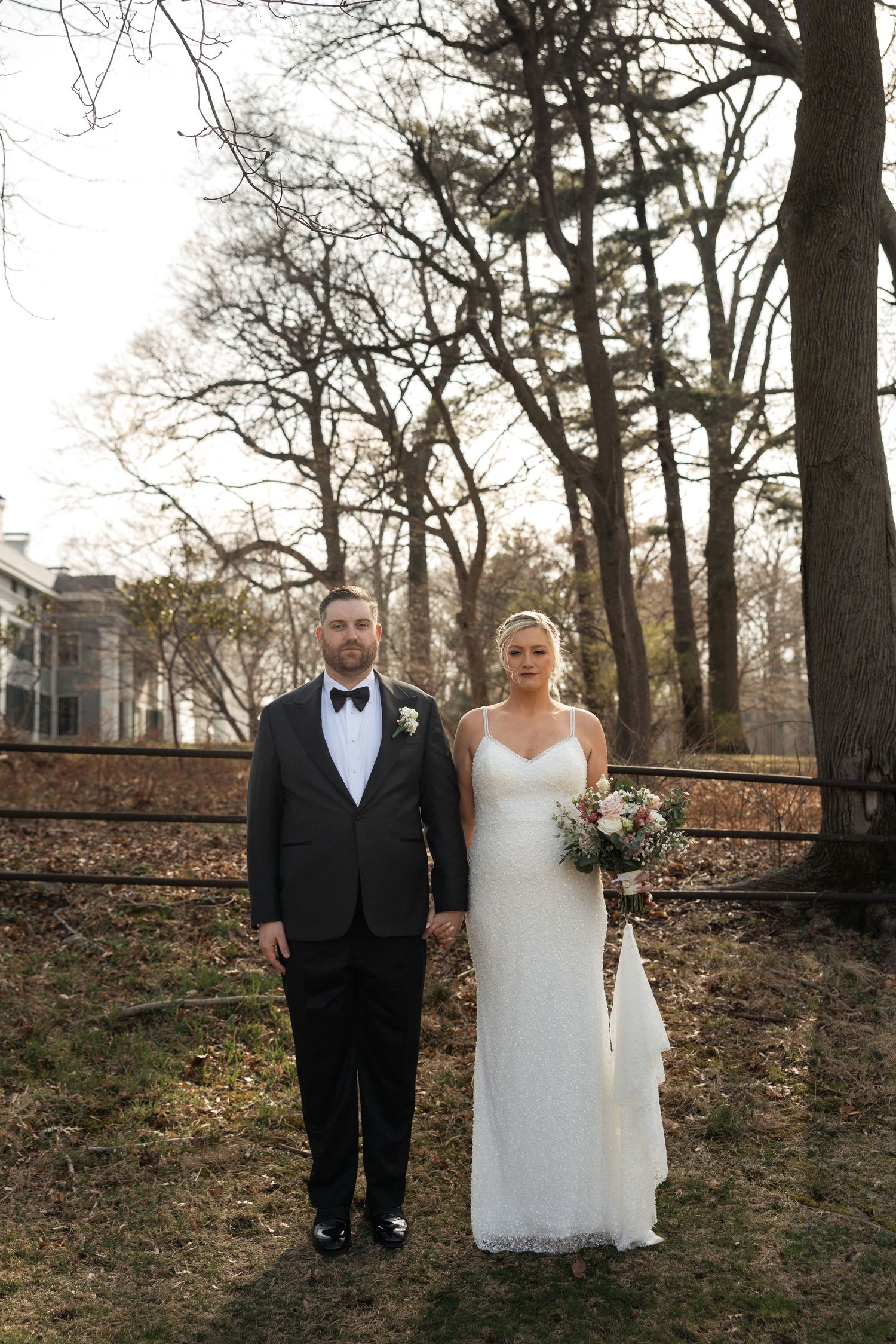 Bride and groom holding hands outdoors in winter, she in white gown with bouquet, he in black tuxedo with bow tie.