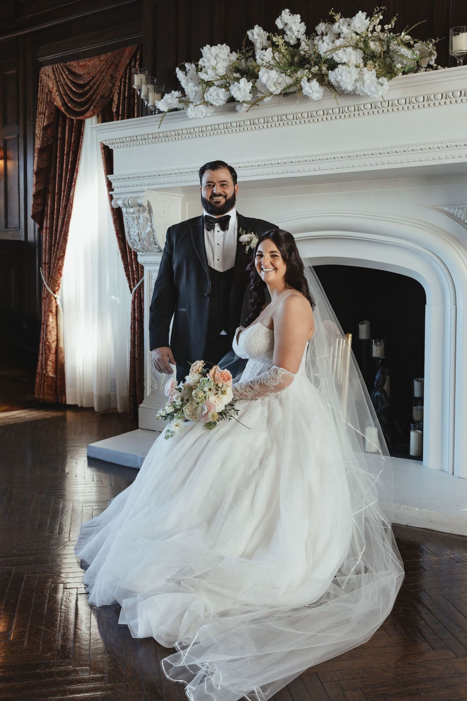 Bride in white ballgown holding bouquet poses with groom in tuxedo before elegant white fireplace adorned with flowers.