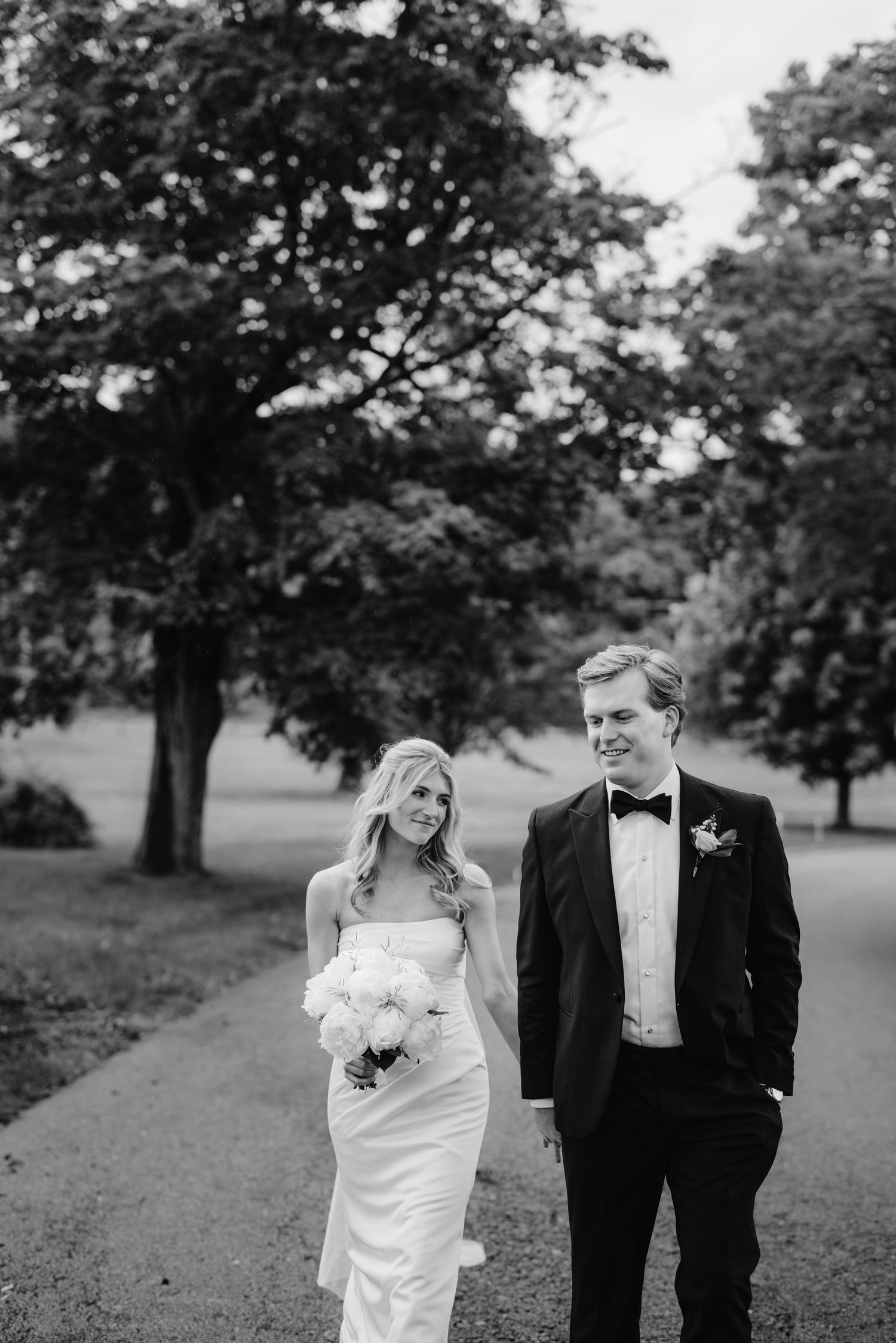 A black and white wedding photo series showing a couple walking and posing together in a park setting with trees.