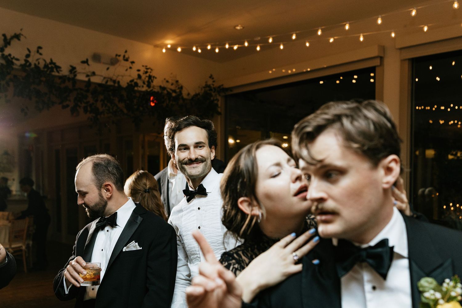 A group of wedding guests dancing and celebrating together under string lights at an evening reception.