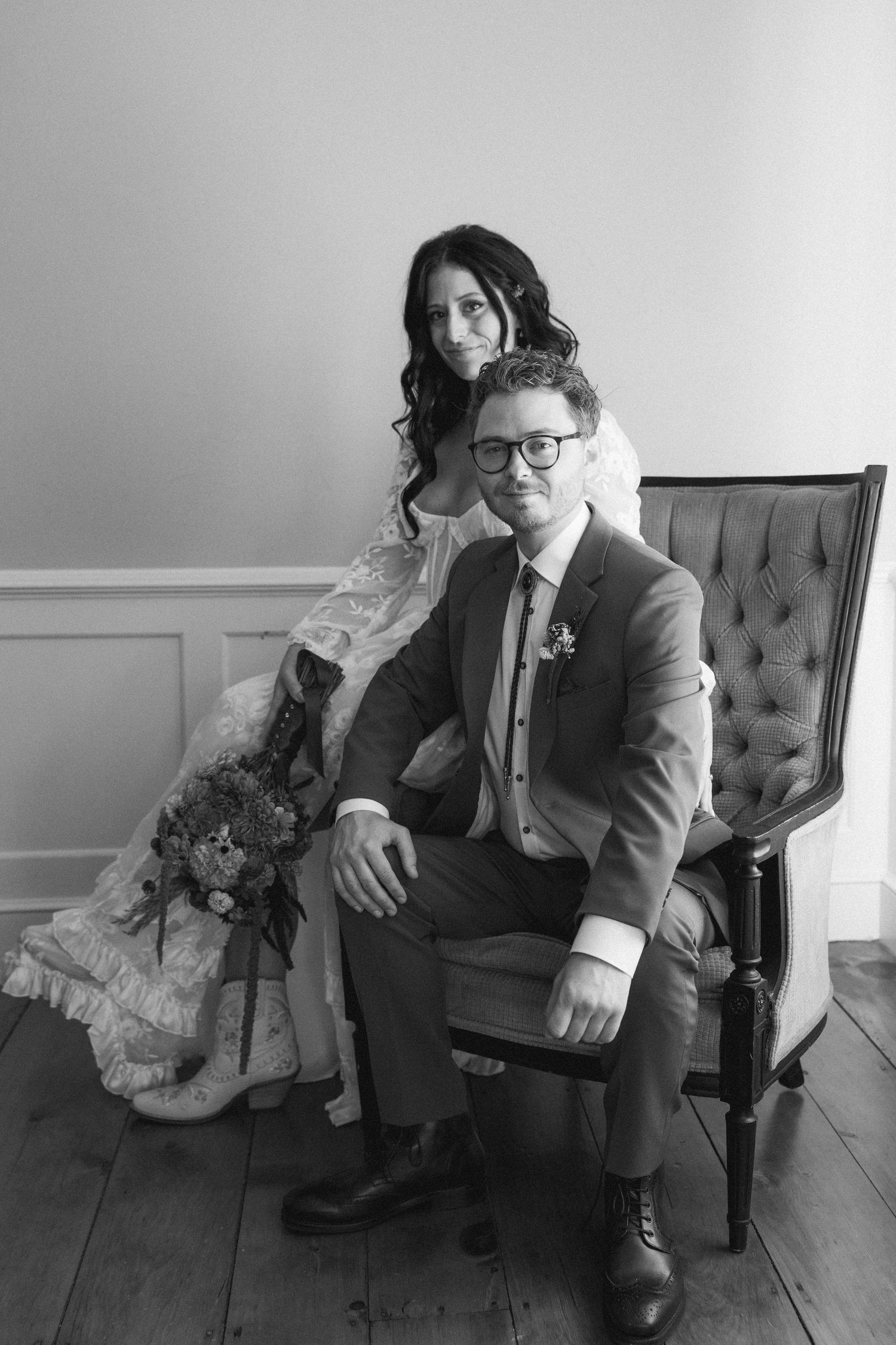 Black and white wedding portrait of a couple posing together on an elegant tufted chair in a classic studio setting.