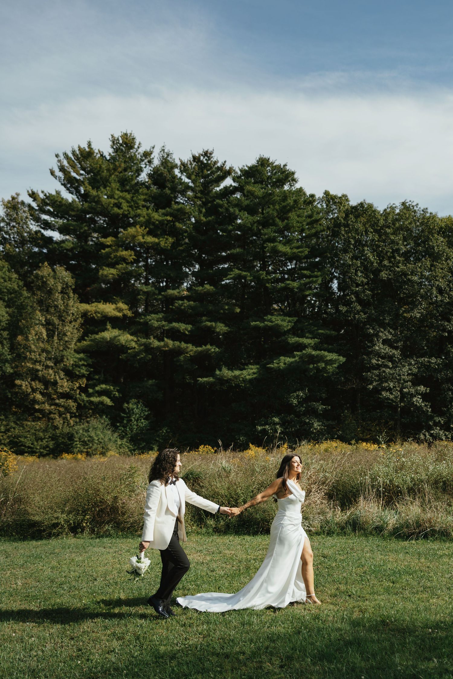 A couple in wedding attire holds hands while walking through a grassy field with tall pine trees in the background.
