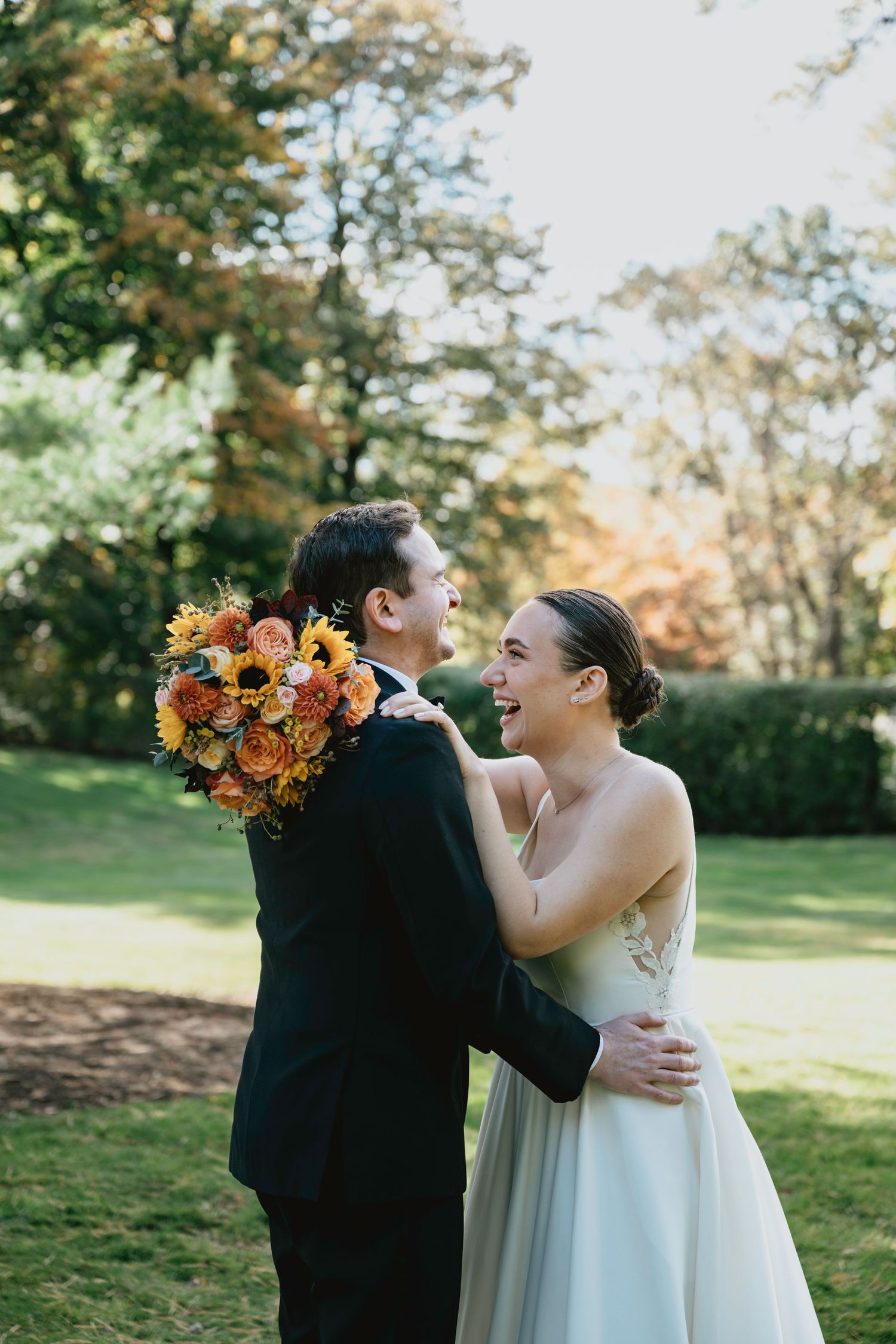 A joyful wedding couple embraces outdoors in autumn with vibrant orange bouquet against green foliage.