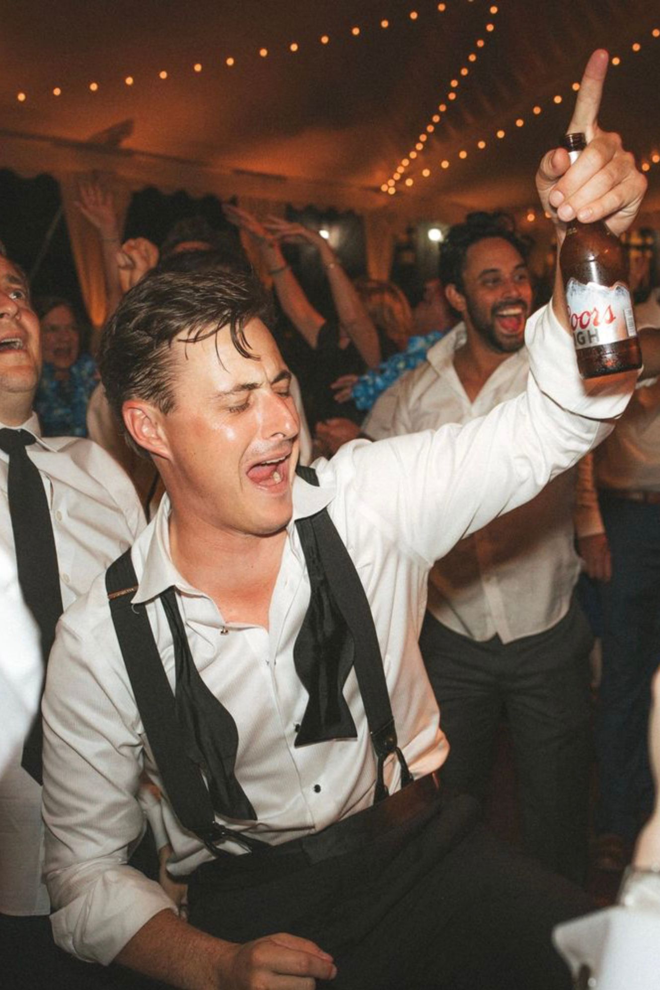Man in suspenders raises Coors beer bottle while dancing at a lively wedding reception party under string lights.