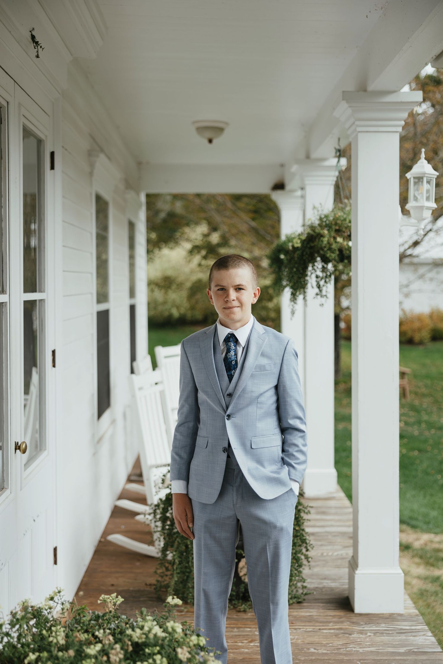 A person in a light gray suit poses thoughtfully along a white columned porch walkway of an elegant venue.