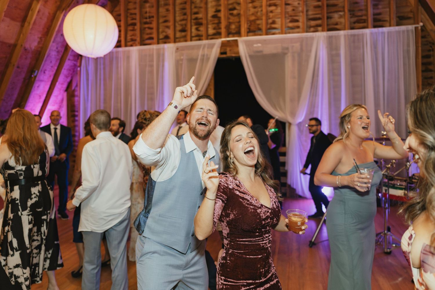 Wedding reception guests dance and celebrate with raised arms under soft purple lighting and white decor.