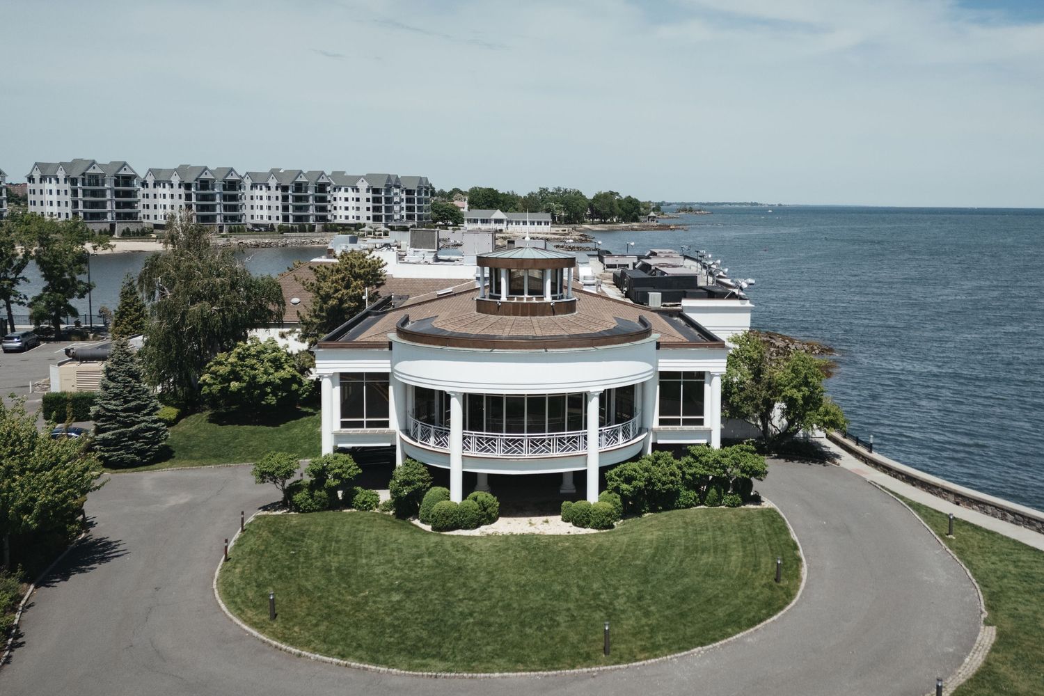 Aerial view of a elegant circular waterfront building with wrap-around porch overlooking a calm bay with marina nearby.