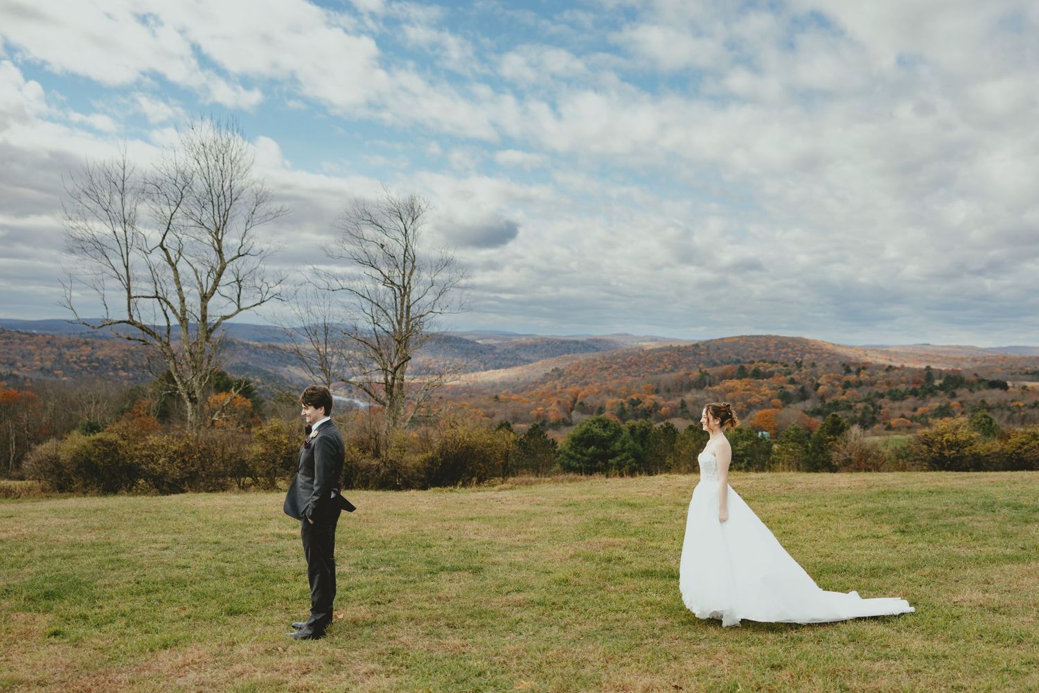 Two people in wedding attire stand apart in a scenic mountain meadow with autumn colors and cloudy skies.