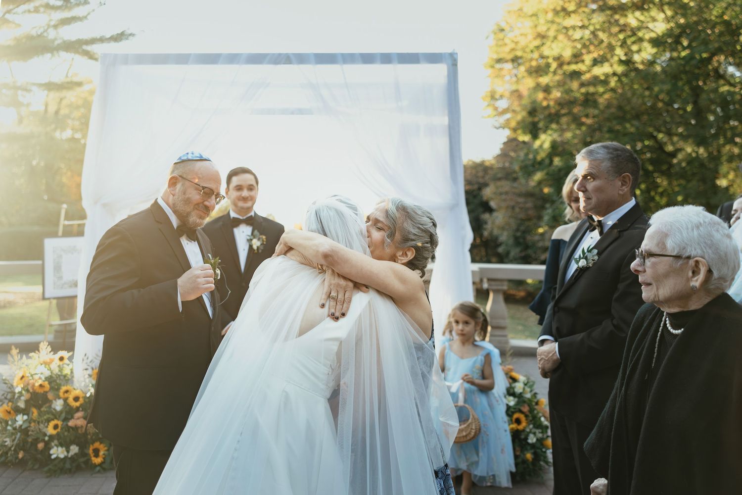 Wedding party gathers outdoors for joyful group photos in a garden setting during golden hour.