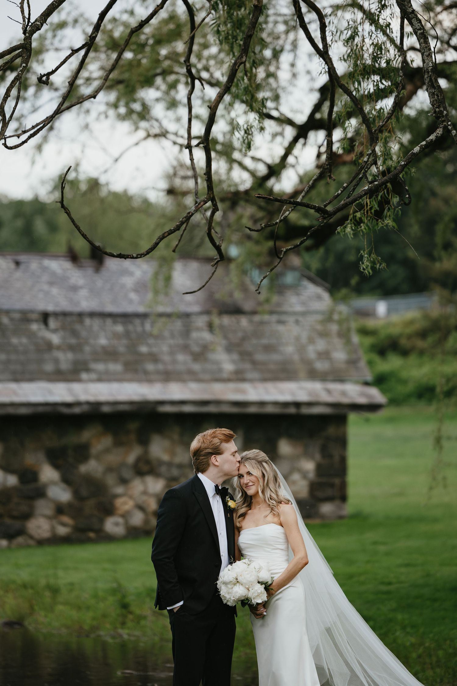 Romantic wedding couple shares intimate moment by stone building and pond under willow branches on overcast day.