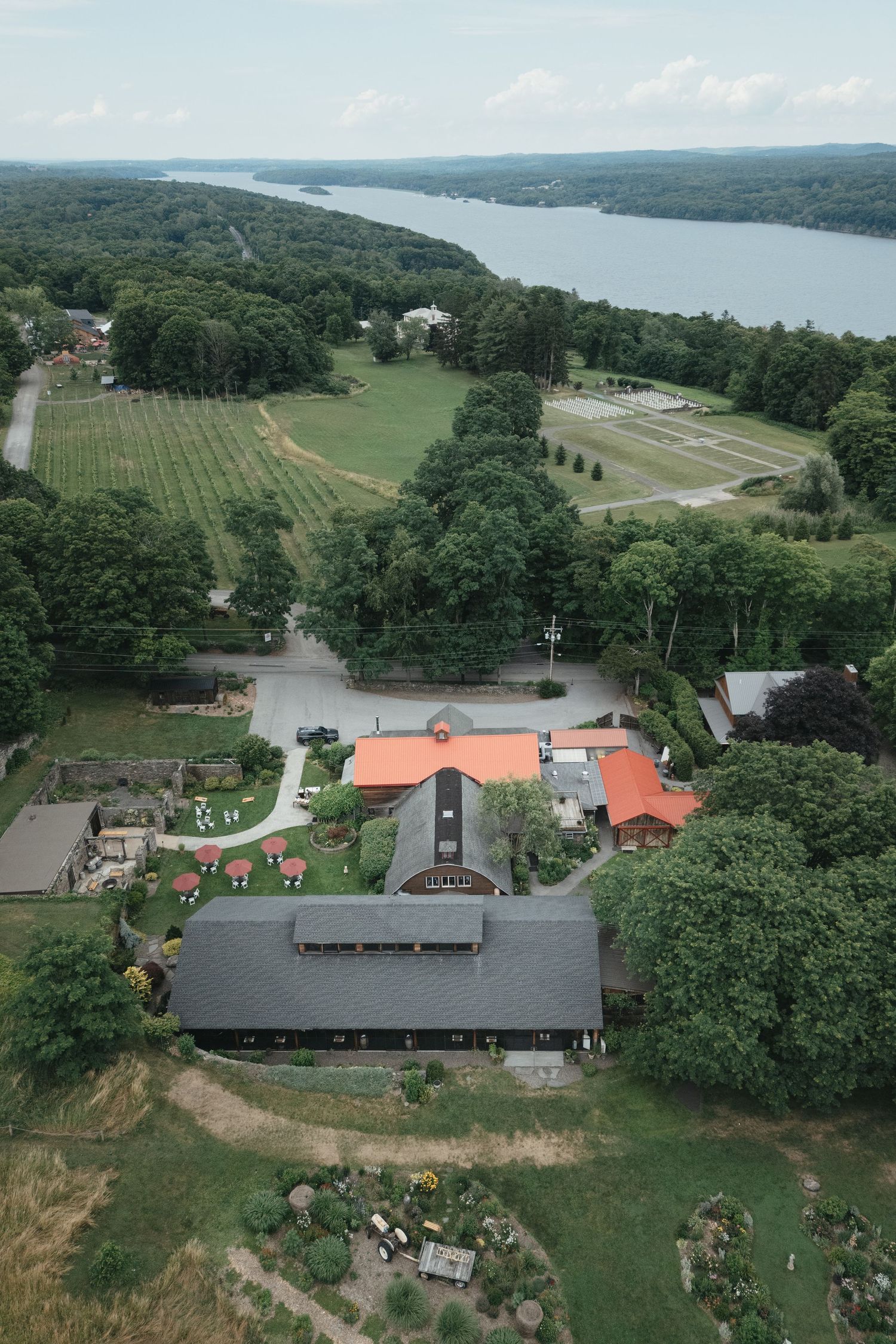 Aerial view of a rural estate with red-roofed buildings, lush green fields, and a serene river in the background.