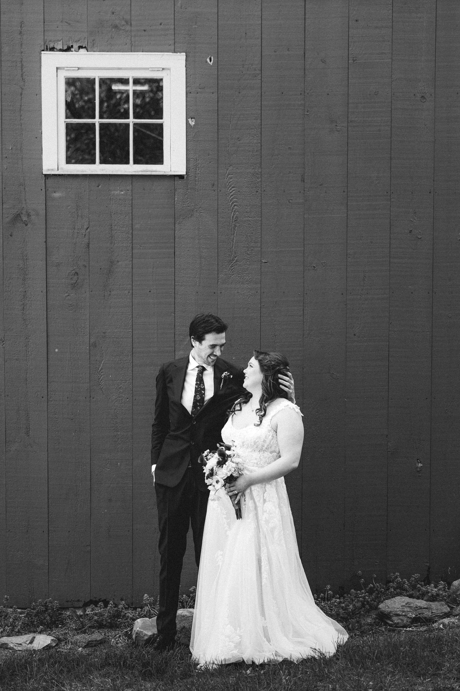 Black and white wedding photograph showing a couple embracing against a rustic barn wall.