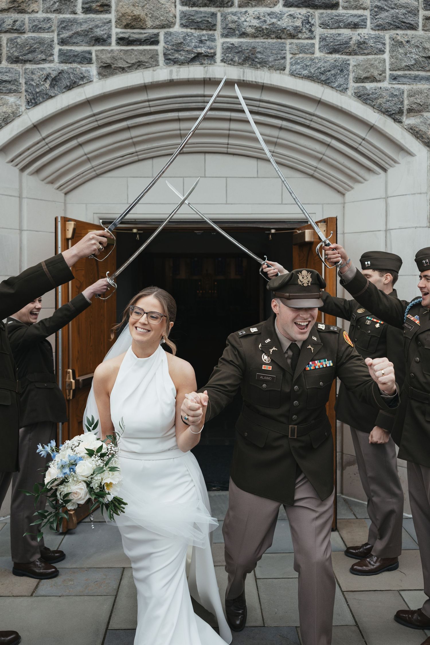Military wedding couple exits church under sword arch, bride in white gown holding bouquet, groom in army uniform.