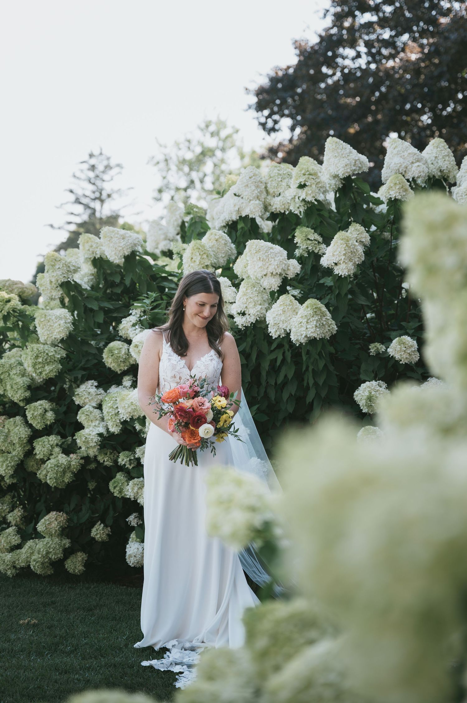 A white floral backdrop frames a bride in a flowing dress holding a vibrant coral and pink bouquet.