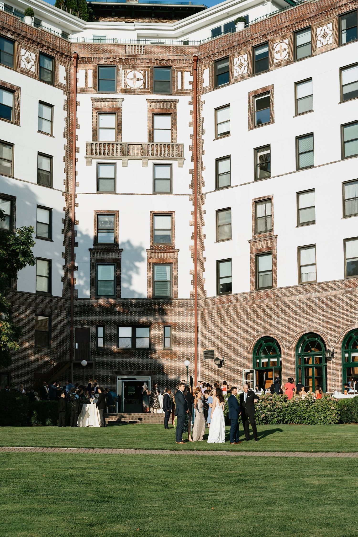 Wedding party gathered on a manicured lawn in front of an elegant brick and white historic apartment building.