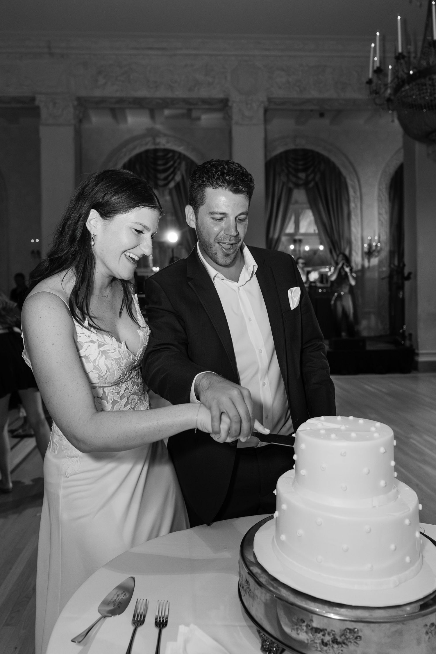 A couple cuts into their elegant white wedding cake together at an ornate reception venue with chandeliers.