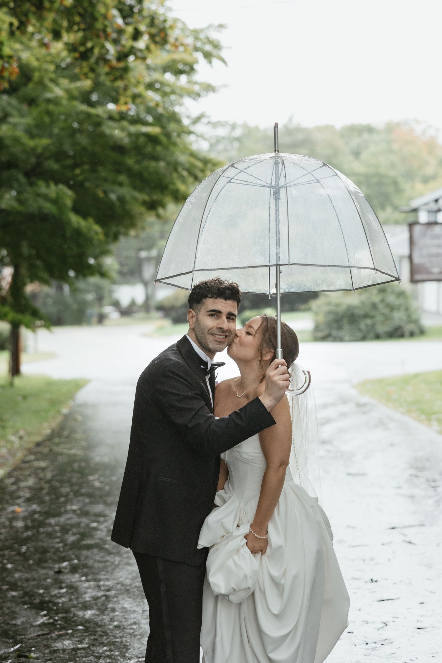 Bride and groom share a kiss under a clear bubble umbrella on a rainy wedding day outdoors.