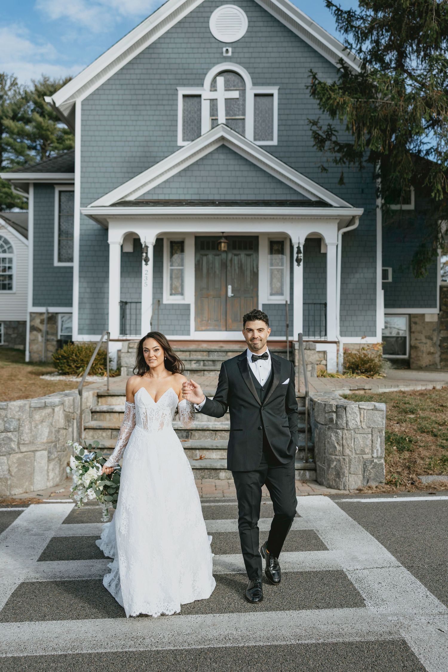 A couple sits on stone steps outside a modern gray house with white trim on their wedding day.