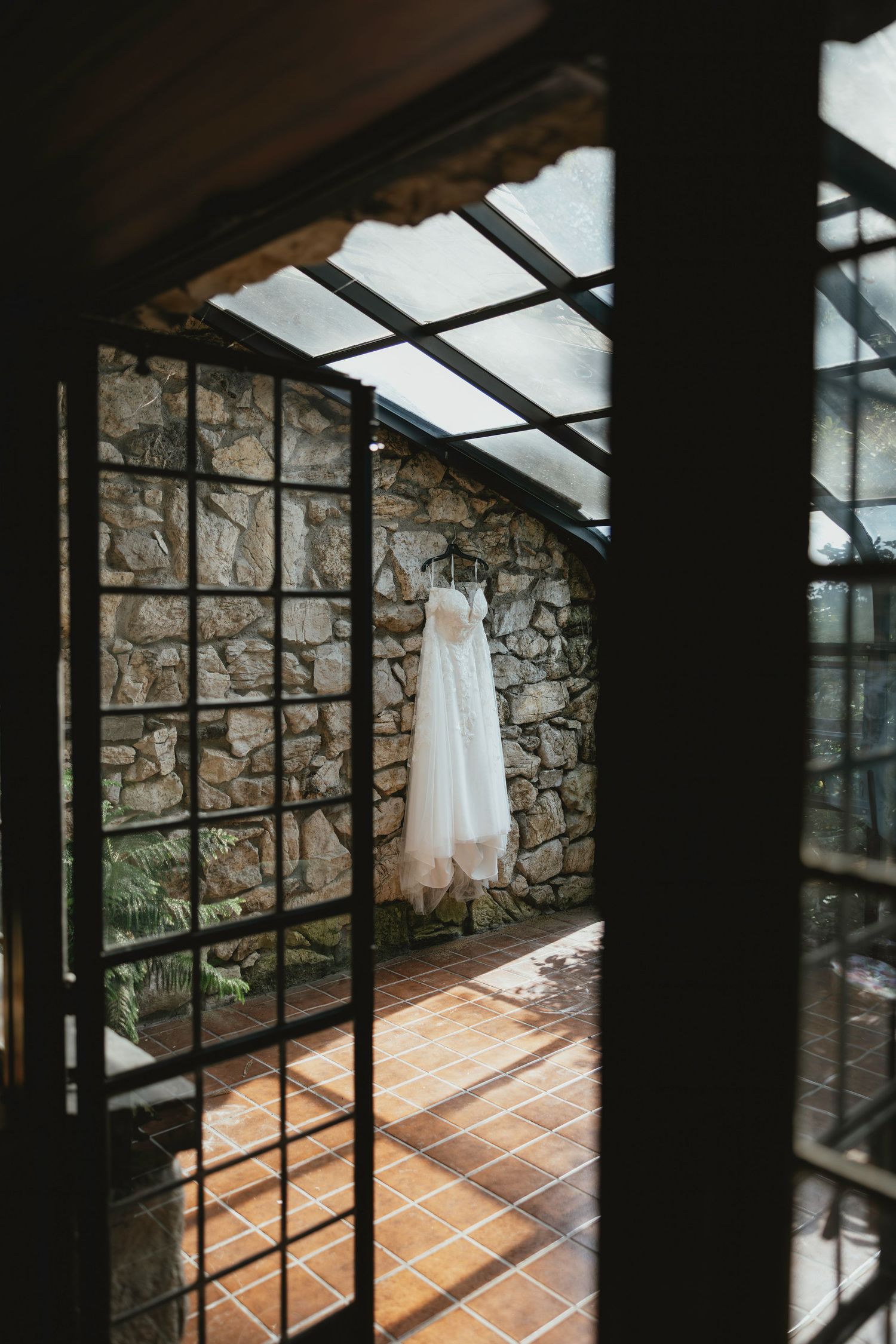 A white wedding dress hangs elegantly in a rustic stone greenhouse with sunlight streaming through glass panels.