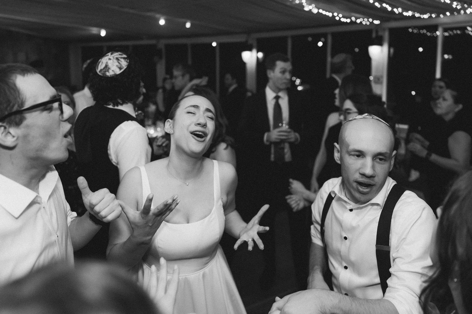Wedding guests dance and celebrate together on a dimly lit dance floor with string lights overhead.