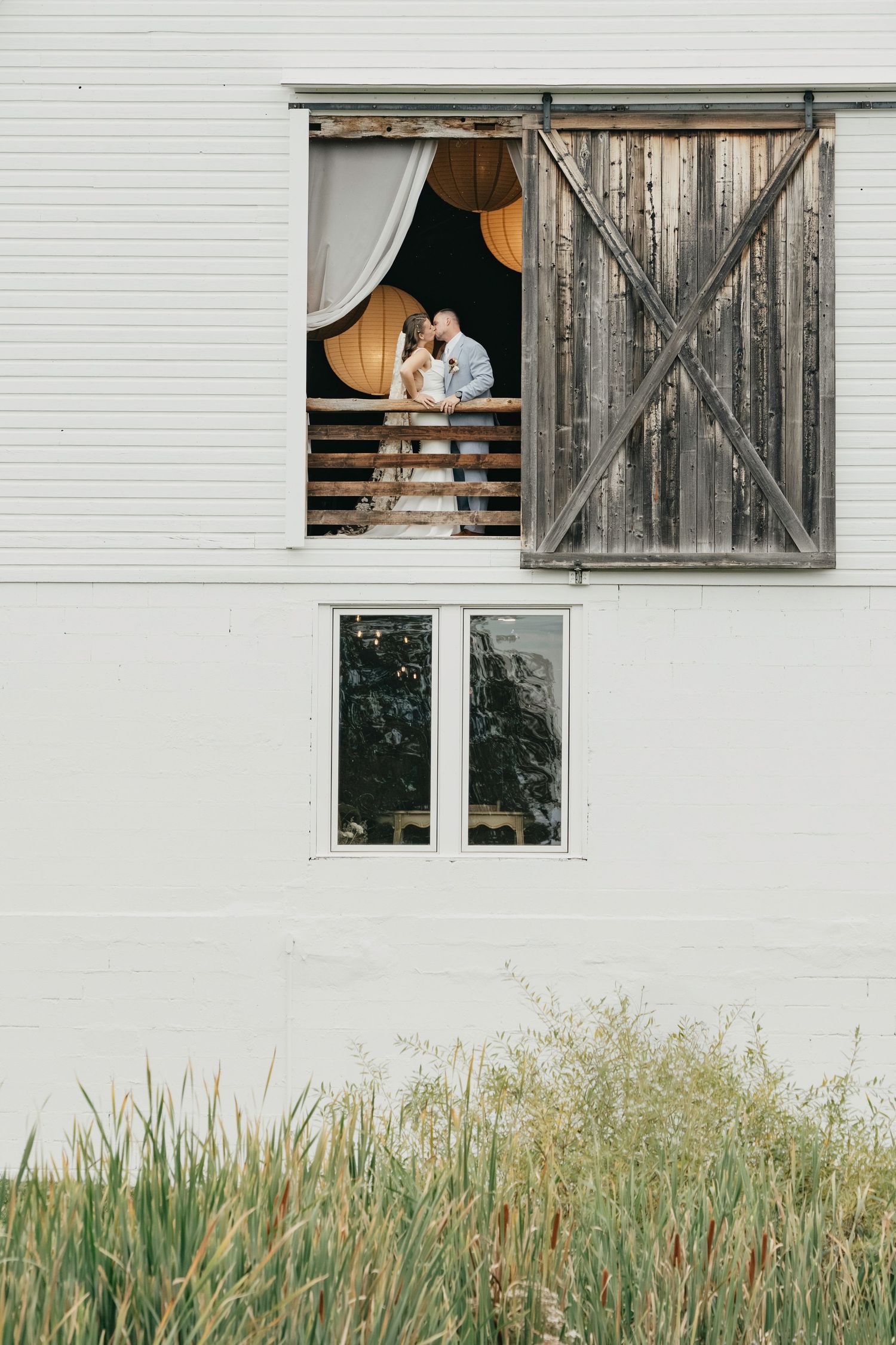 A white barn with rustic wooden doors features tall grass growing along its foundation.
