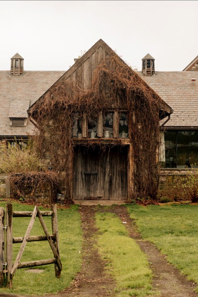 Rustic abandoned barn covered in dried vines with wooden doors, stone walls, and a grass pathway leading to the entrance.