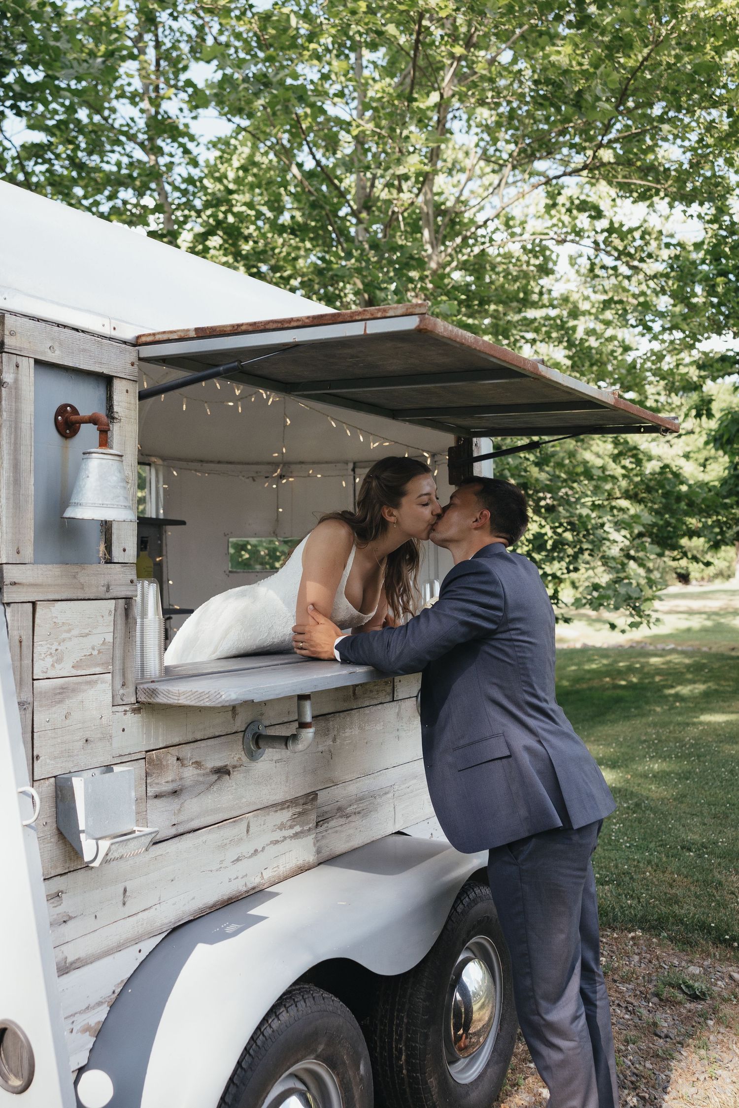 Bride and groom share a romantic kiss at a rustic food truck window outdoors on their wedding day.
