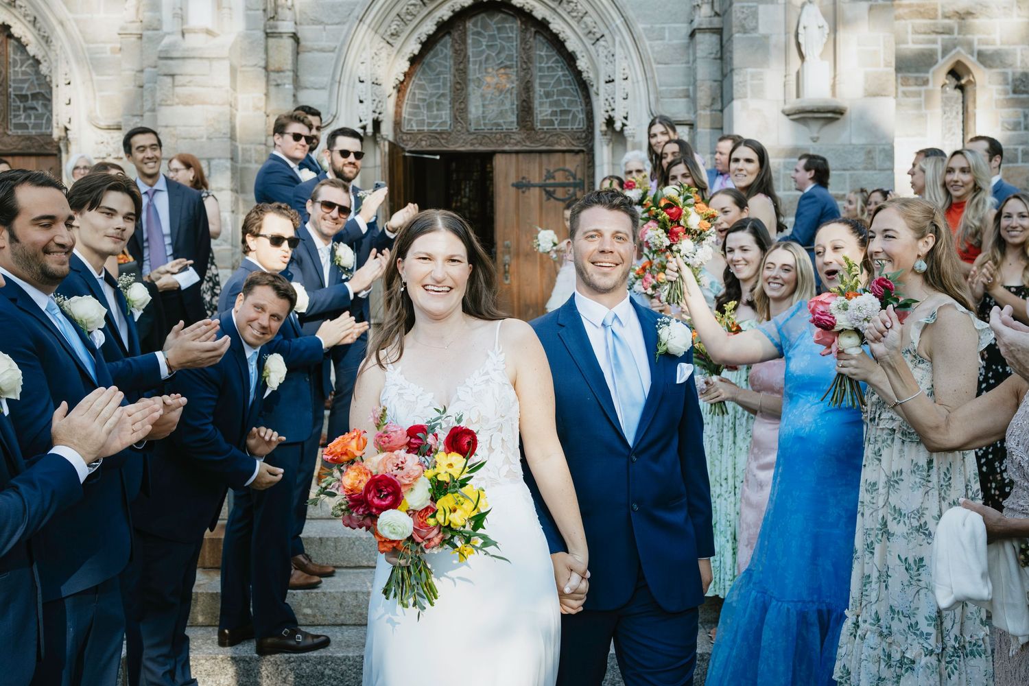 Wedding party celebrates outside historic stone church with colorful bouquets and formal attire.