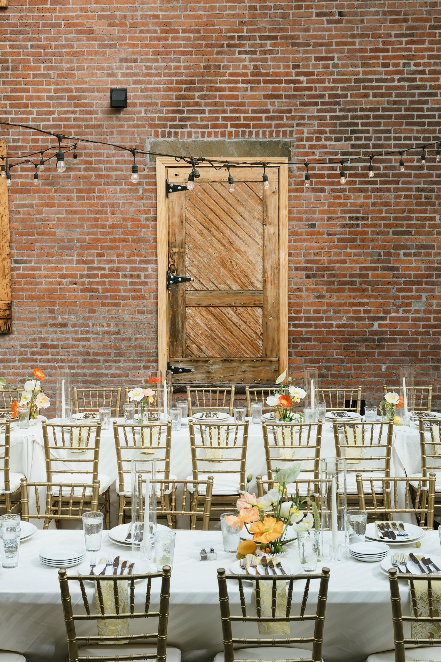 Elegant wedding reception tables set with white linens and gold chiavari chairs against rustic brick wall and wooden doors.