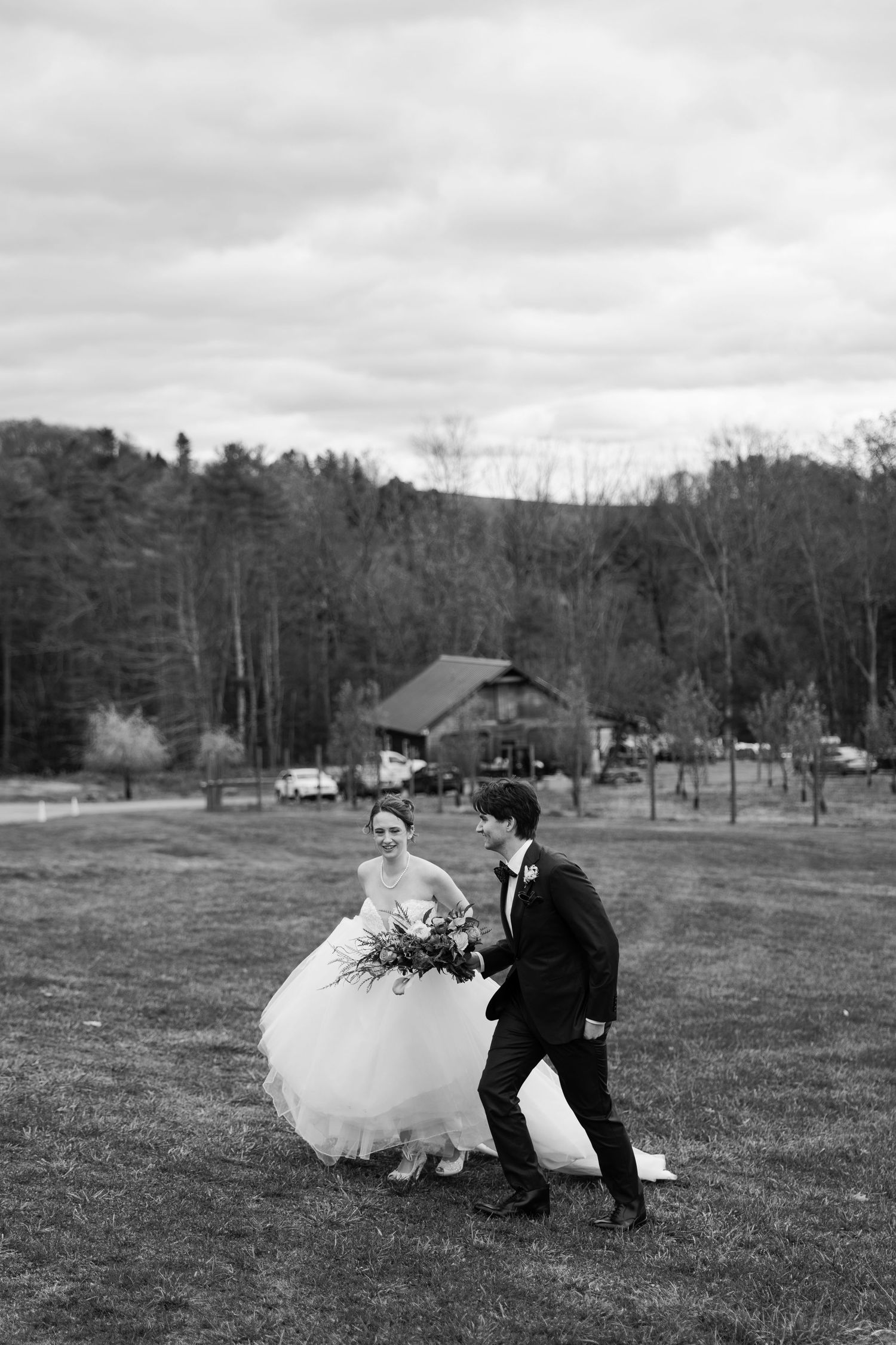 Black and white photo of newlyweds running playfully across a grassy field with cabin in background.