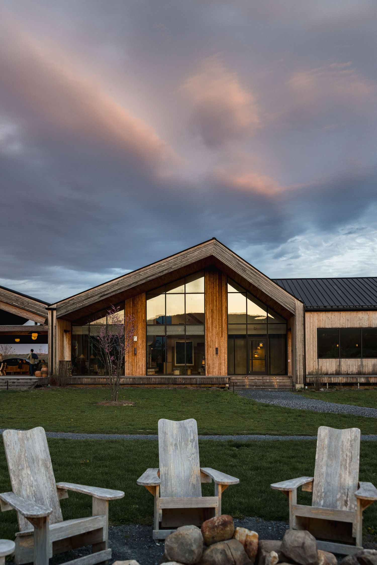 Rustic lodge with wooden gable facade, large windows reflecting sunset, Adirondack chairs around stone fire pit at dusk.